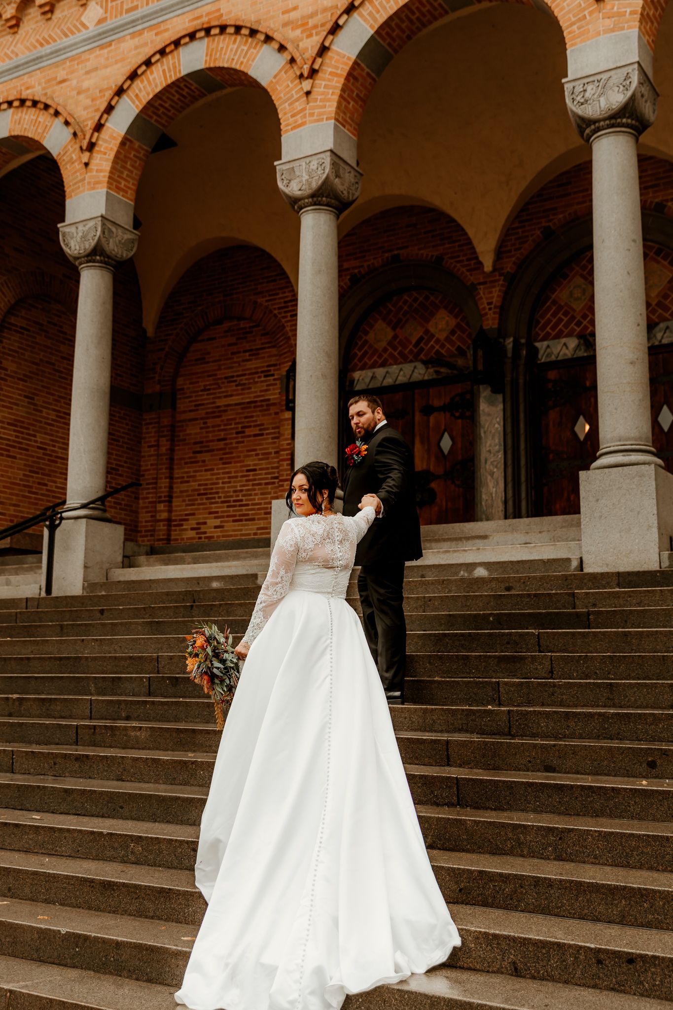 Bride in a flowing white gown holding a bouquet leads her groom up stone steps to a brick church with arched columns.