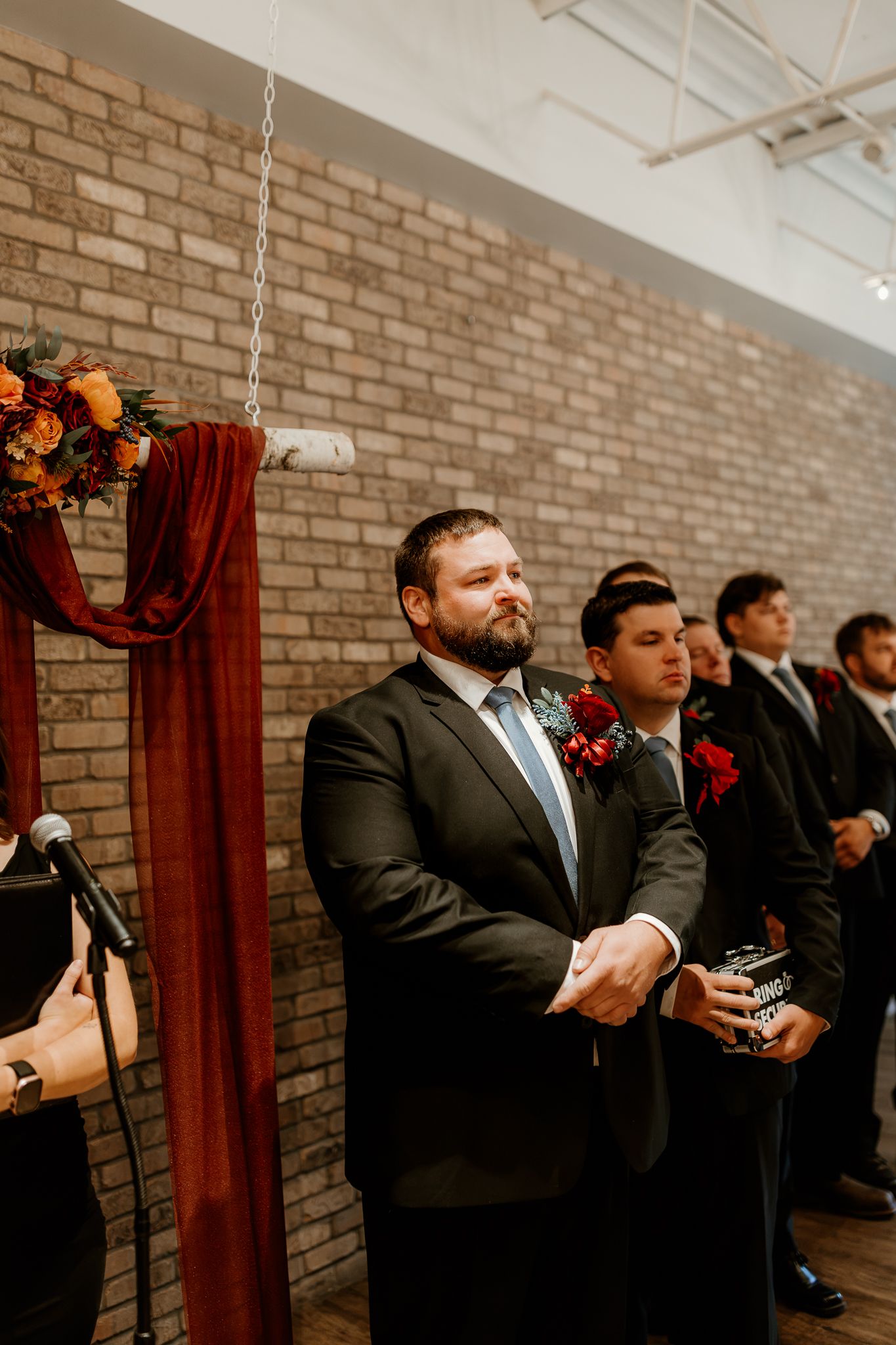 Groomsmen in dark suits with burgundy boutonnieres stand at a wedding ceremony beside a brick wall and rust-colored fabric drapery.