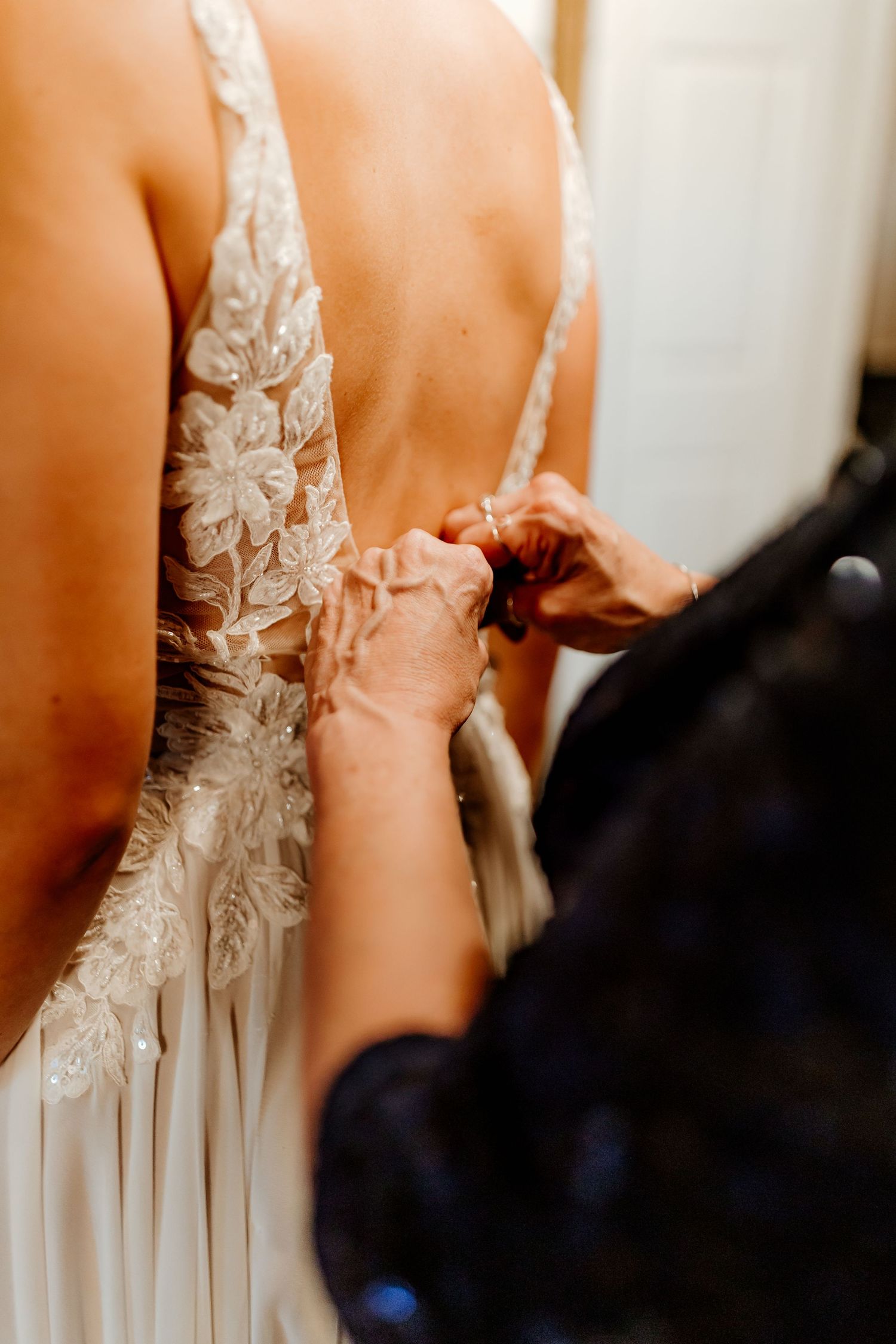 Hands button the back of a white lace wedding gown with floral appliqué details as the bride gets ready for her ceremony.