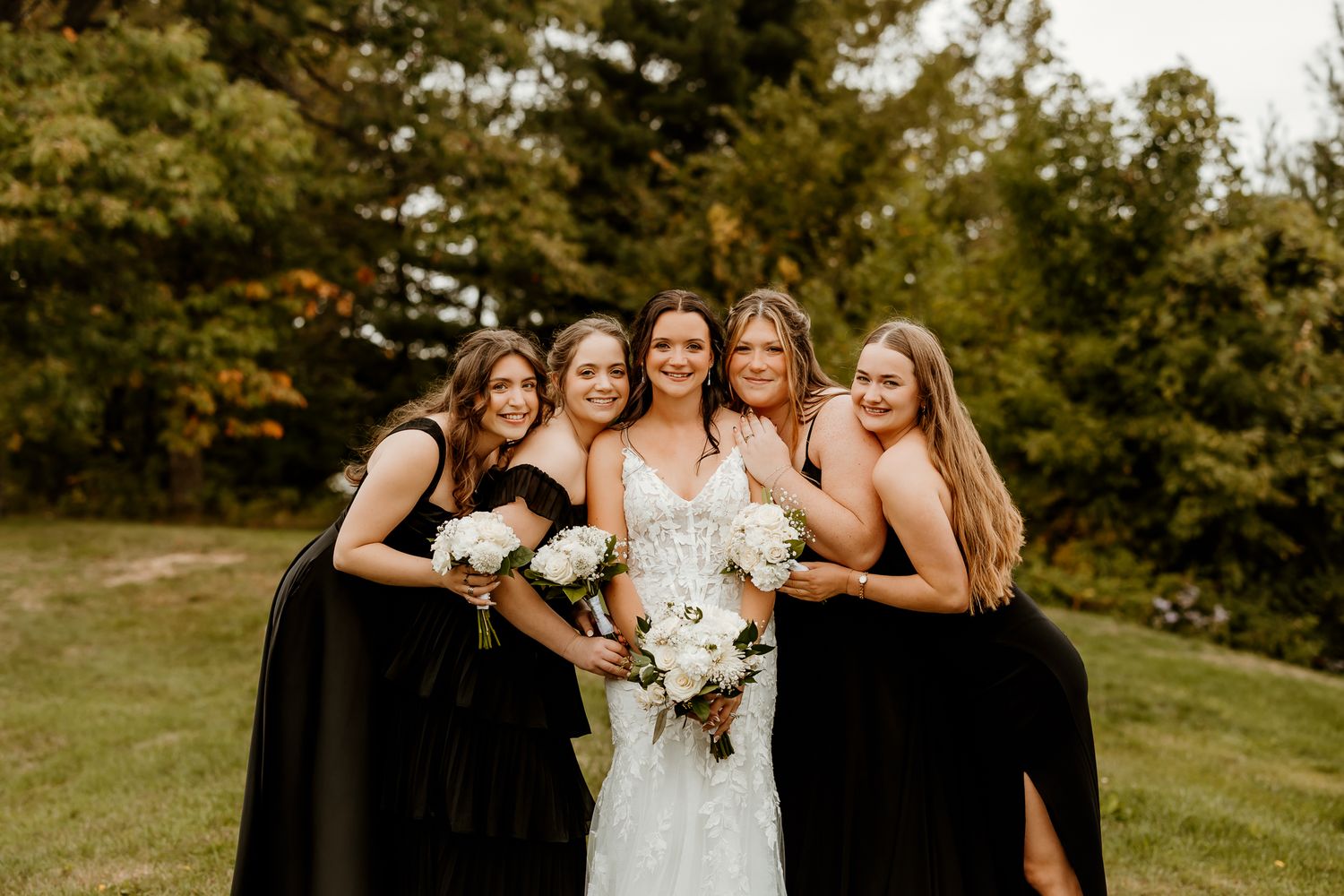 Bride in a white lace gown laughs with four bridesmaids in black dresses holding white bouquets on a grassy lawn near trees.
