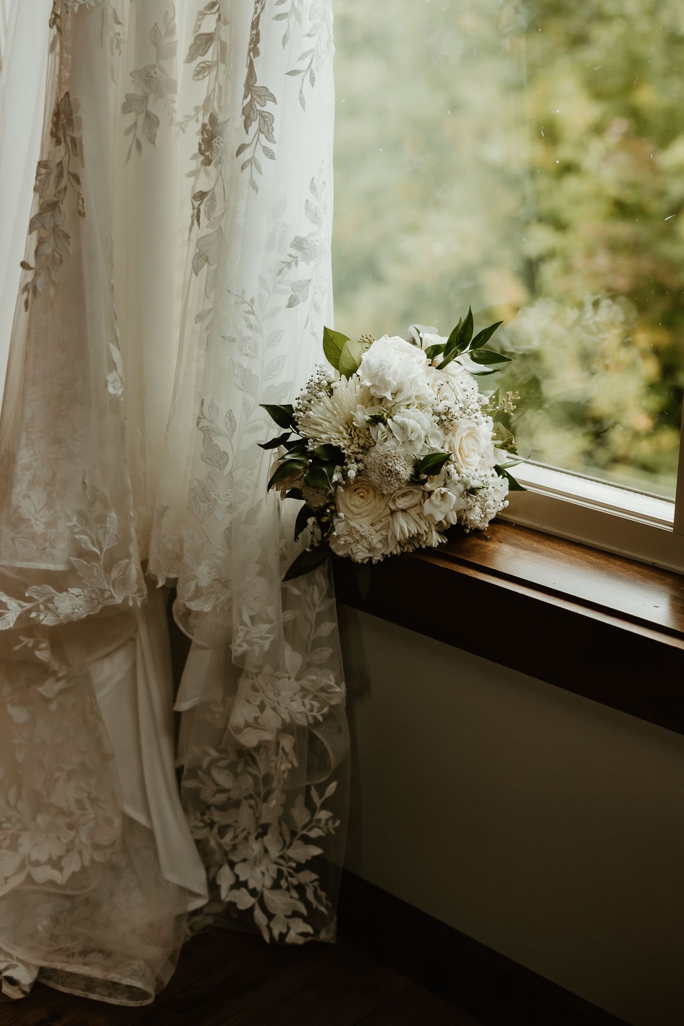 White floral wedding bouquet with greenery resting on a wooden windowsill beside a lace wedding gown and a sunlit window.