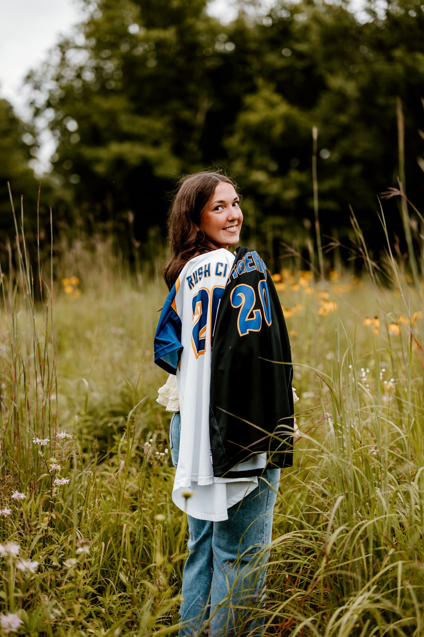 Smiling teen girl in Rush City jersey #20 standing in tall wildflower field with lush green trees behind her.