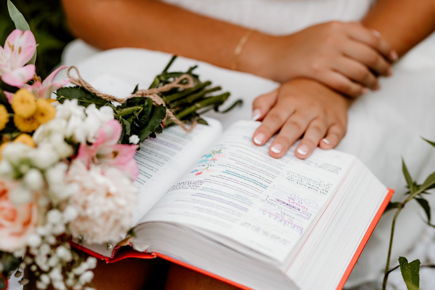 Bride holding open Bible with highlighted verses beside colorful wedding bouquet of pink and yellow flowers.
