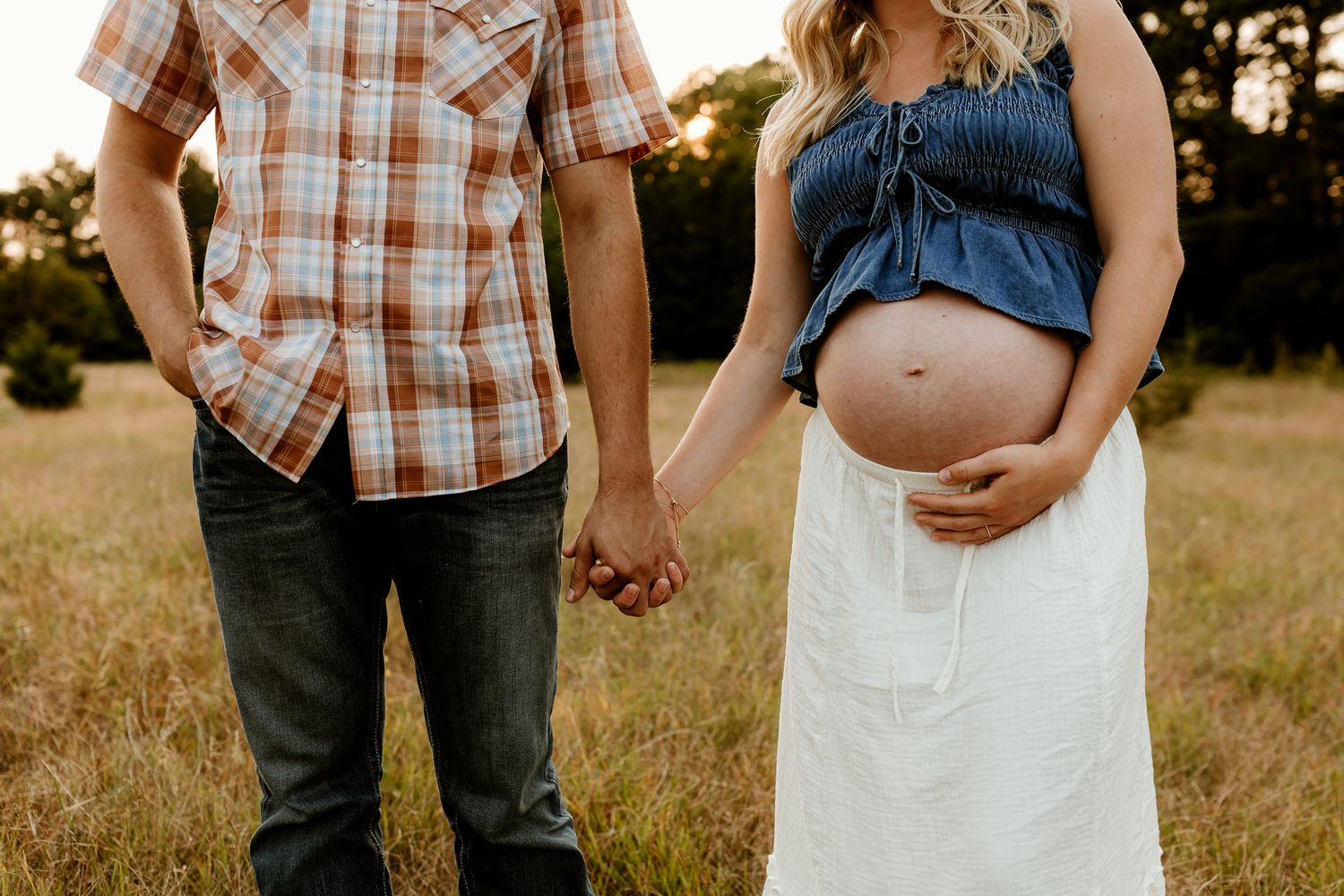 Pregnant couple holding hands in a golden field, woman in denim top and white skirt showing baby bump.