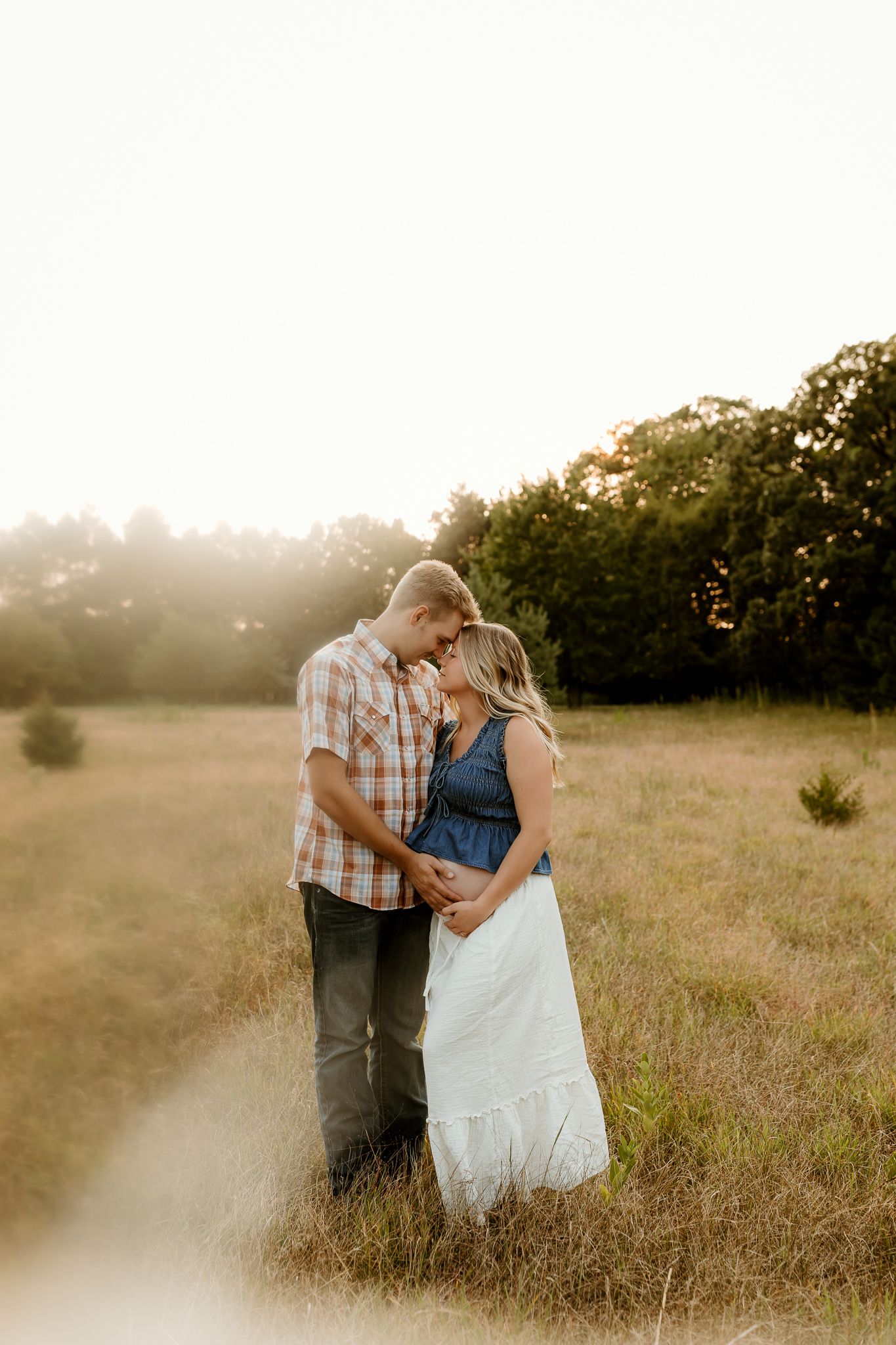Couple embracing in golden hour sunlight in a grassy field, woman in denim and white dress, man in plaid shirt.