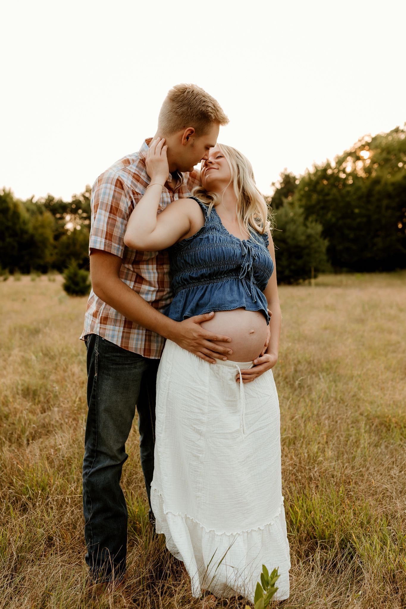 Couple sharing a tender moment in a golden field, man kissing pregnant woman's forehead while cradling her baby bump.