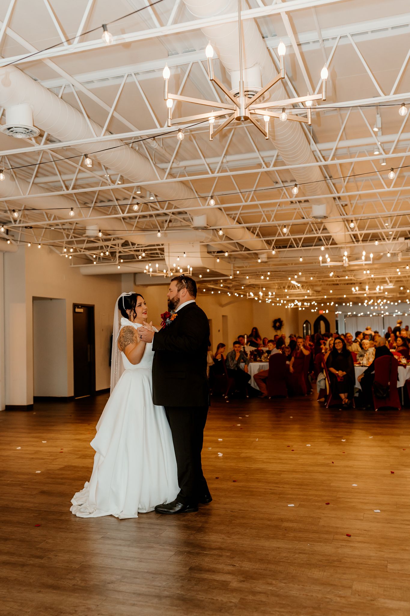 Bride in a white gown and groom in a dark suit share a first dance on a wooden floor under string lights as wedding guests watch.