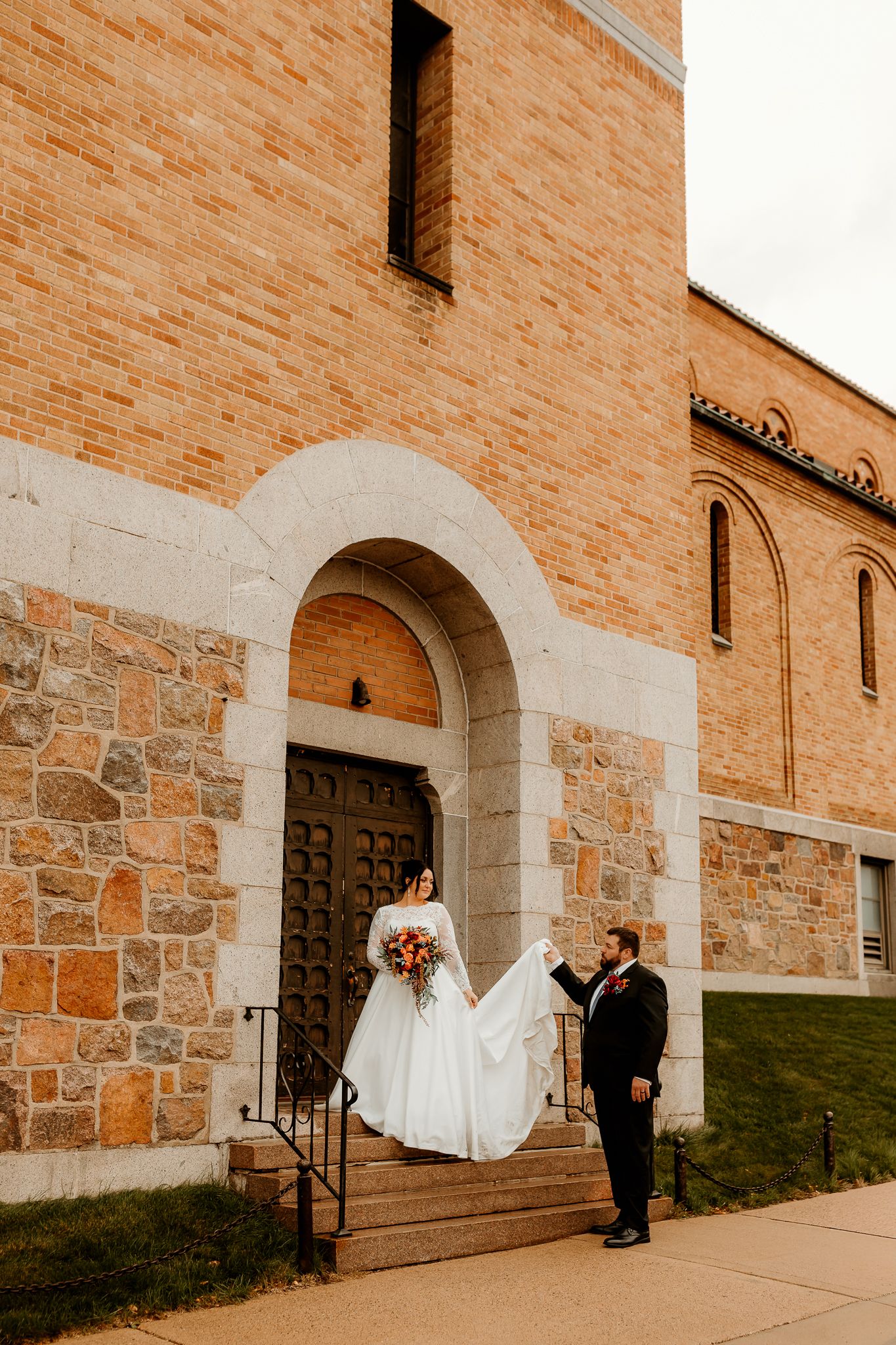 Bride in a white gown holding a bouquet and groom in a dark suit hold her long train on the steps of a brick and stone church.