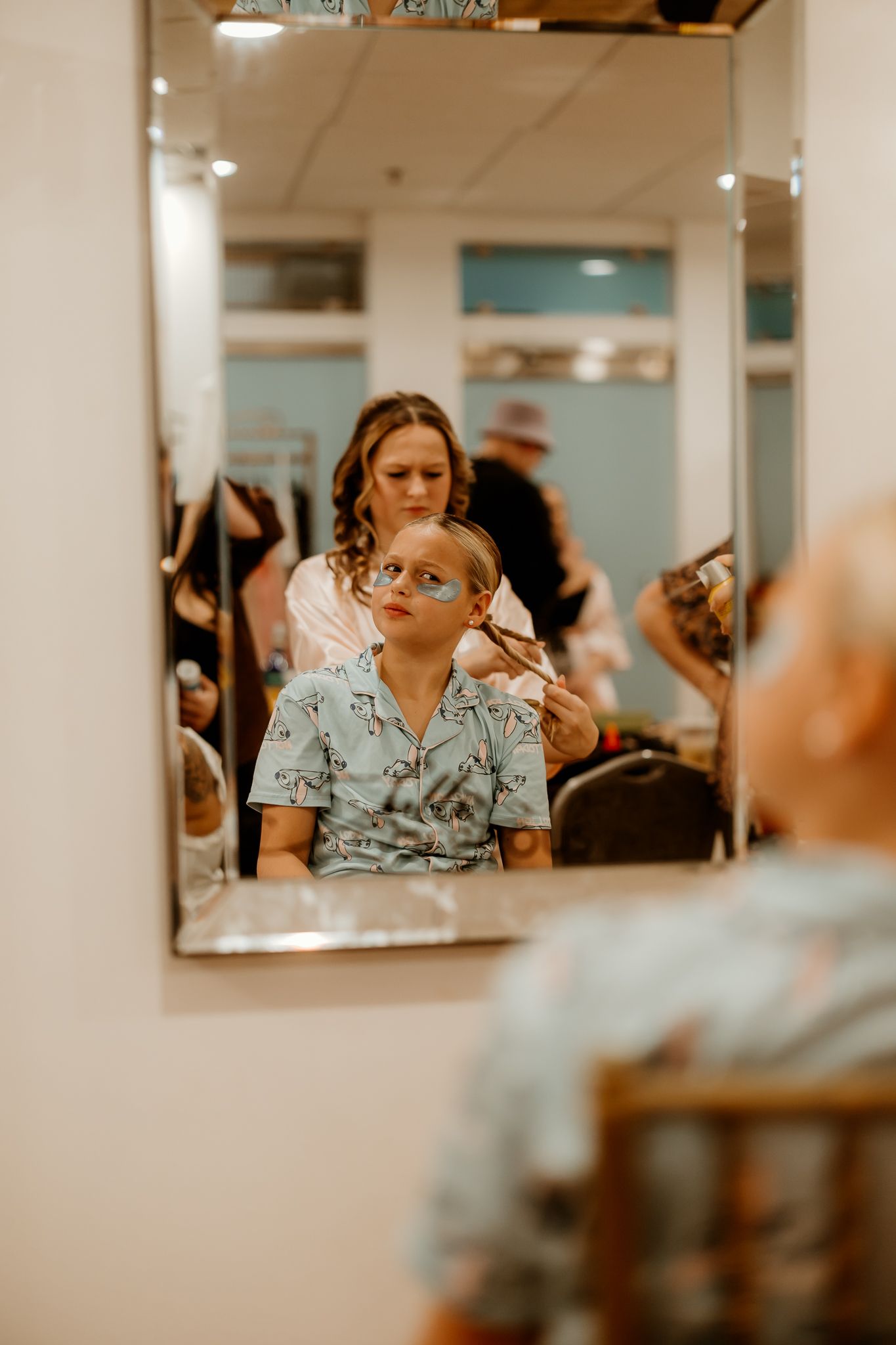Mirror reflection of a young girl in a patterned pajama shirt getting face makeup applied by a woman with curly hair in a bright salon.