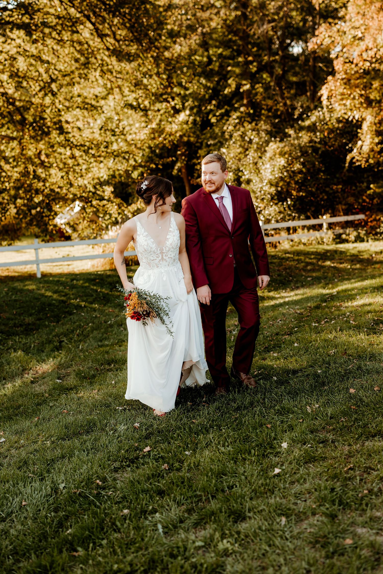 Bride in a white lace wedding gown holding a bouquet walks hand in hand with a groom in a burgundy suit across a sunlit grassy field.