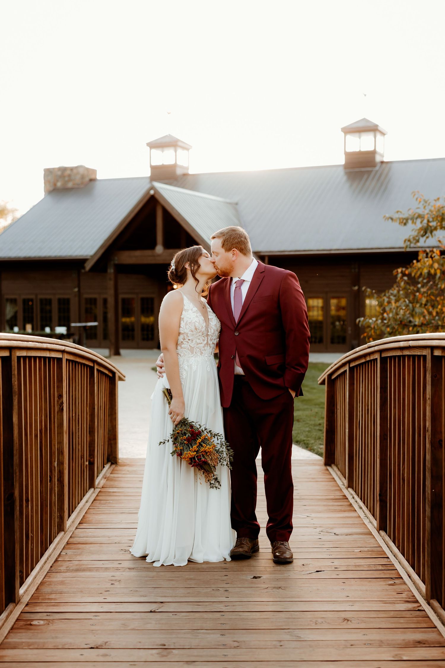 Bride in a lace wedding gown and groom in a burgundy suit kiss on a wooden bridge, holding a bouquet, with a rustic venue behind them.