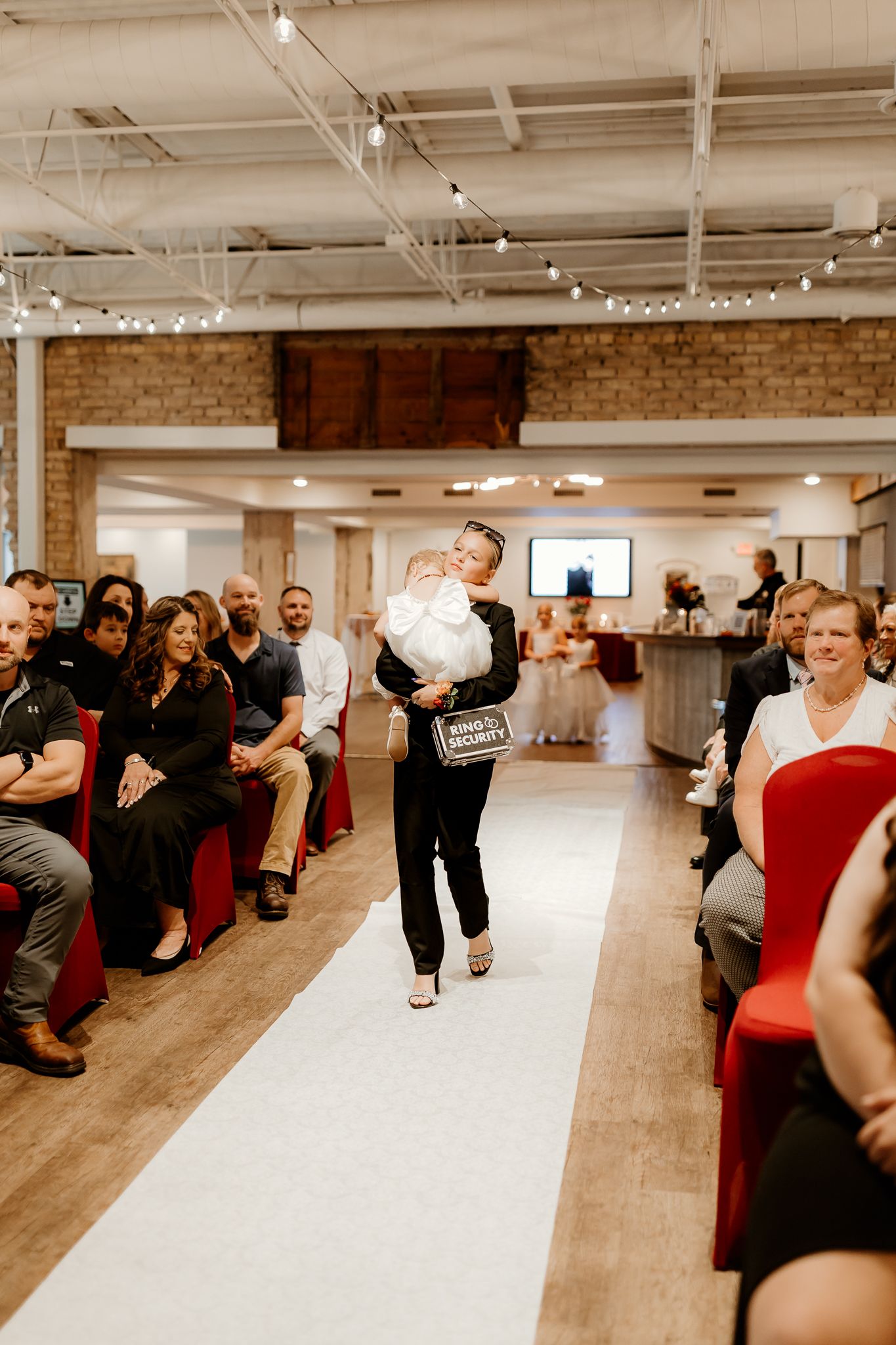 Woman in black carries a baby in white dress and a sign down the white aisle runner as seated wedding guests watch in a brick venue.
