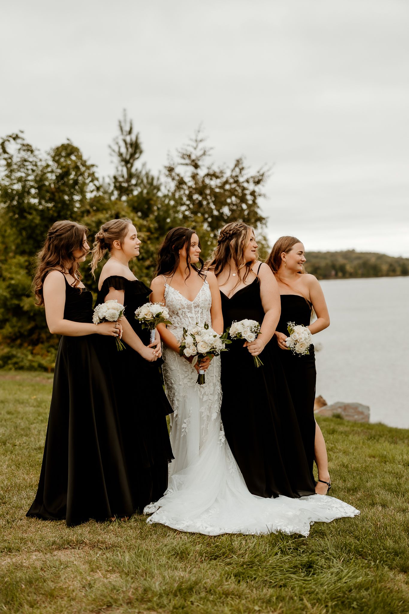Bride in a lace wedding gown poses with four bridesmaids in black dresses holding white bouquets on a grassy lakeside shore.