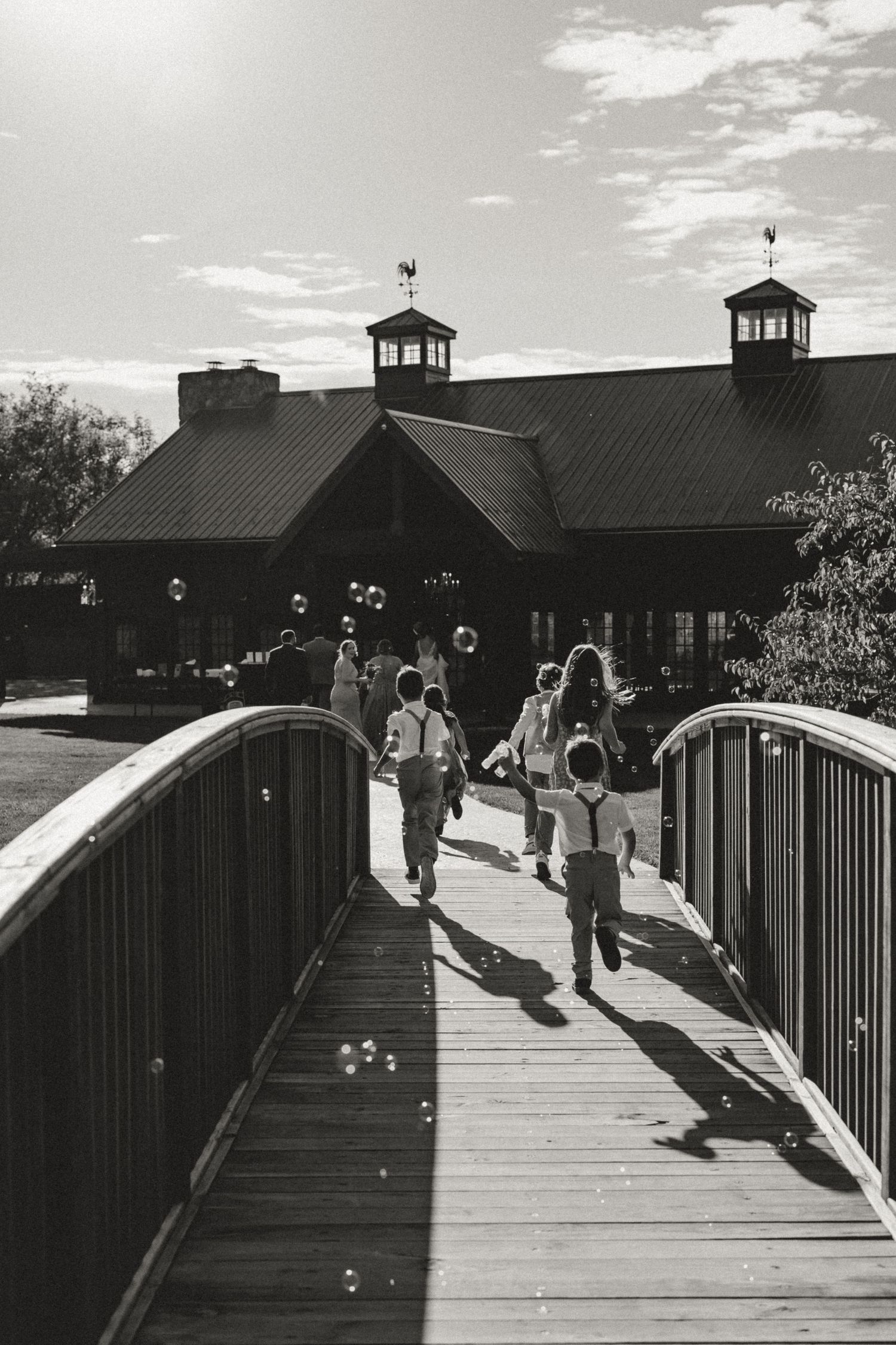 Children run across a wooden bridge in black and white toward a rustic lodge with cupolas, as soap bubbles float through the sunlit air.