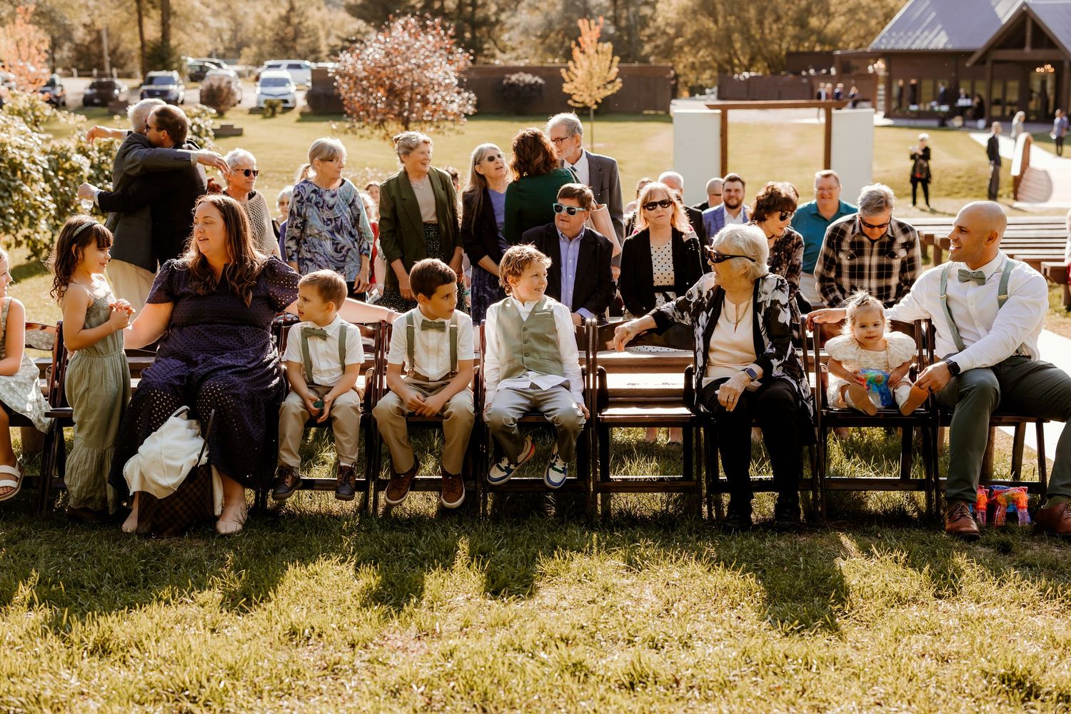 Wedding guests in formal attire sit on wooden benches and stand on a sunny lawn at an outdoor ceremony with autumn trees in the background.