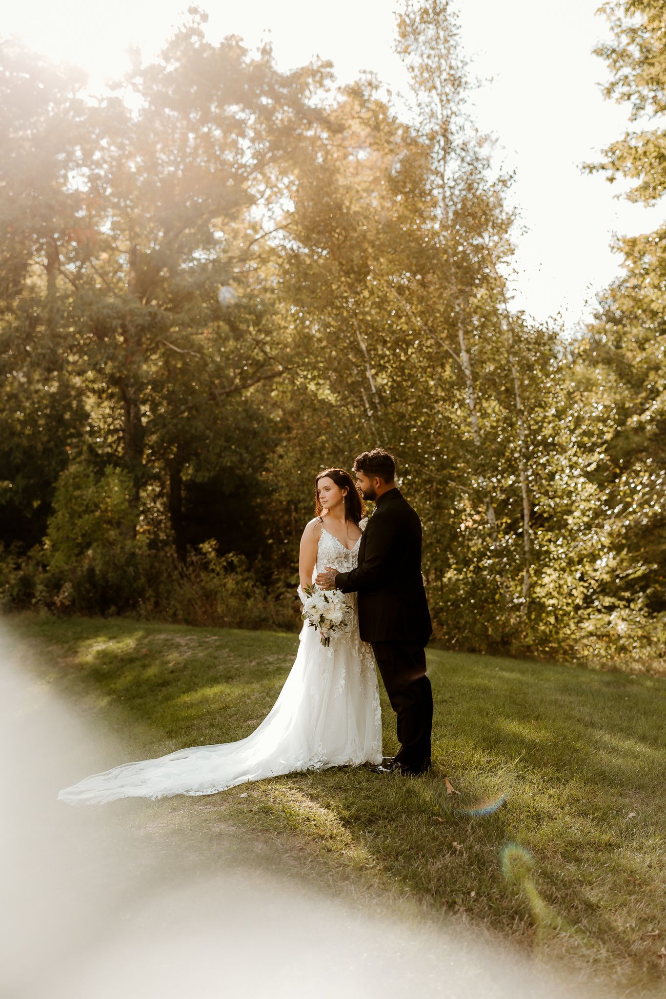 Bride in a lace gown with bouquet and groom in a dark suit embrace on a grassy lawn framed by sunlit autumn trees at an outdoor wedding.