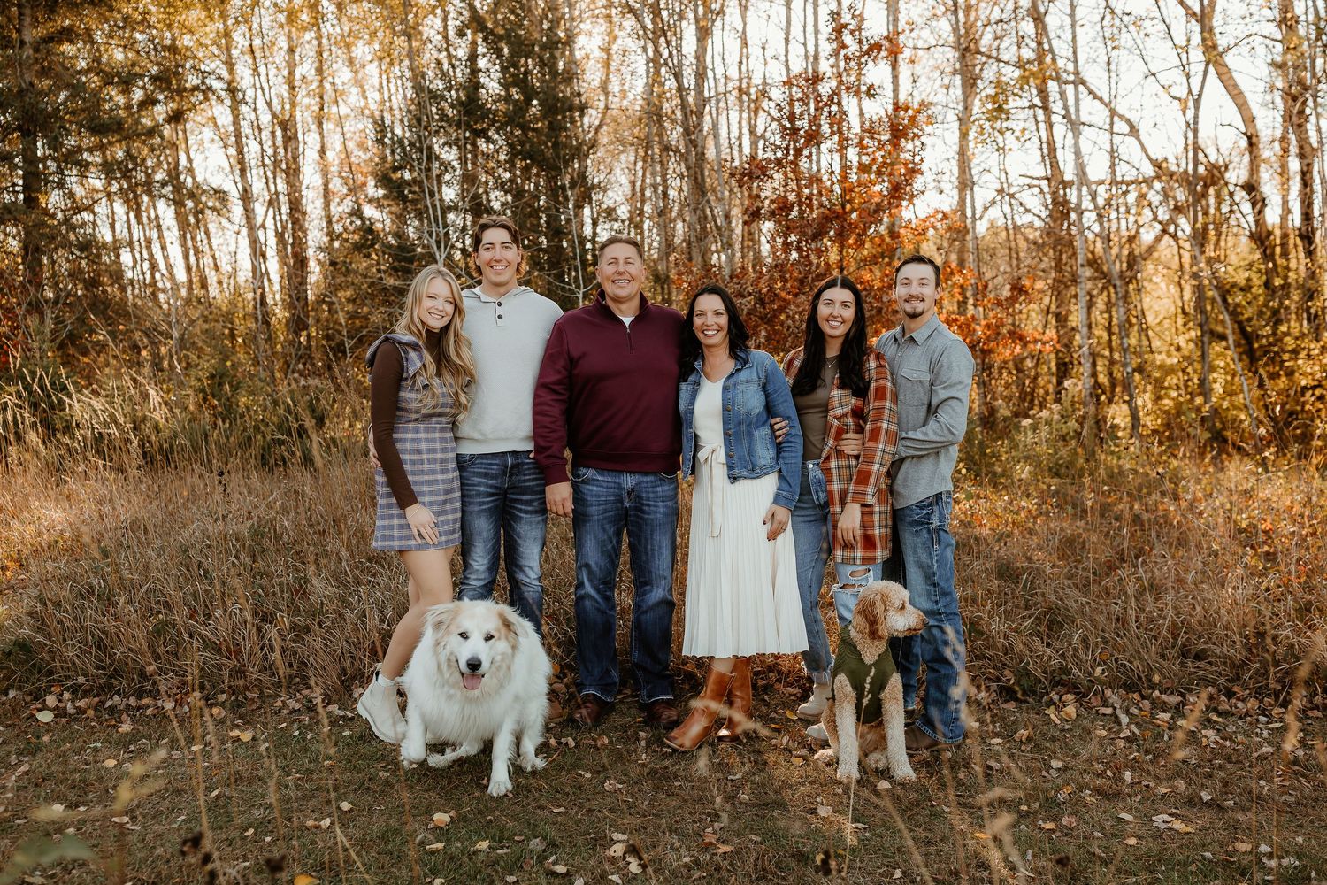 Family of six in casual fall clothing poses outdoors in an autumn forest with a large white dog and a golden dog at their feet.