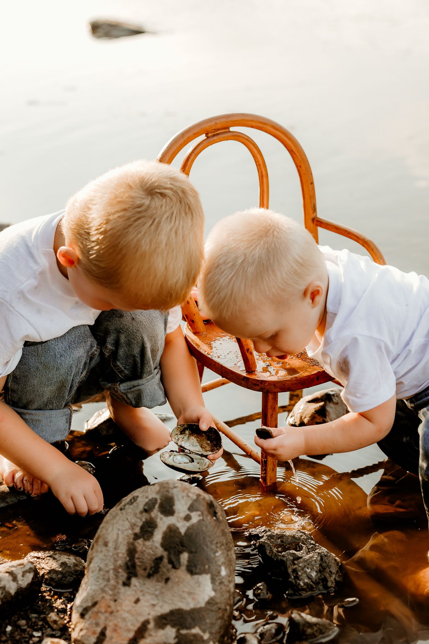 Two young blonde boys in white shirts playing with a wooden chair in shallow water among rocks outdoors.