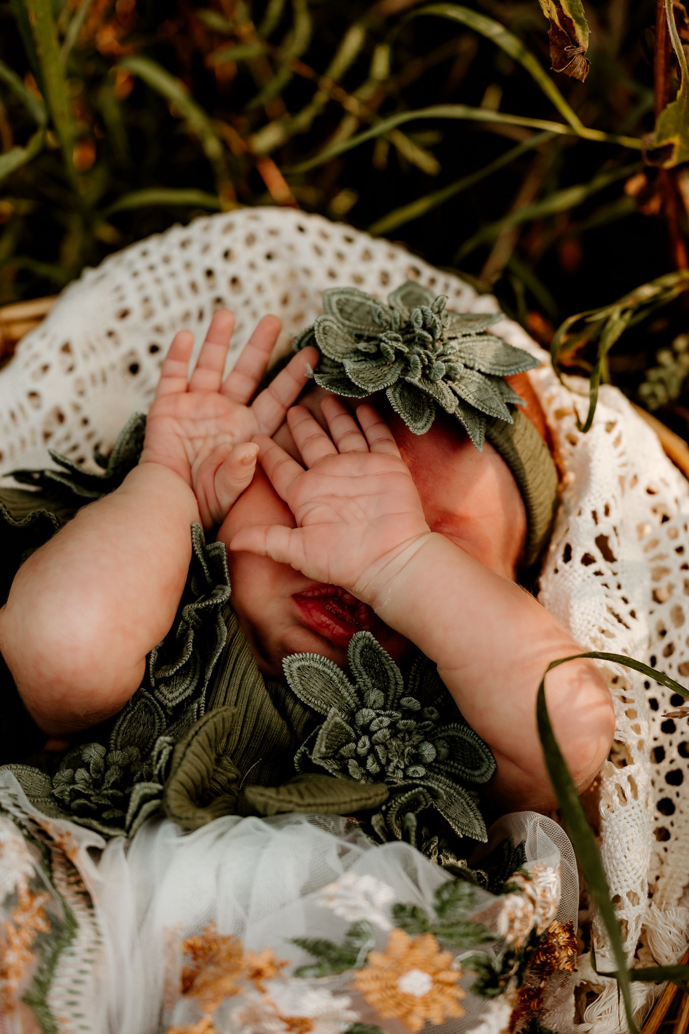 Newborn baby in green floral outfit and hat, hands raised, resting in a wicker basket outdoors.