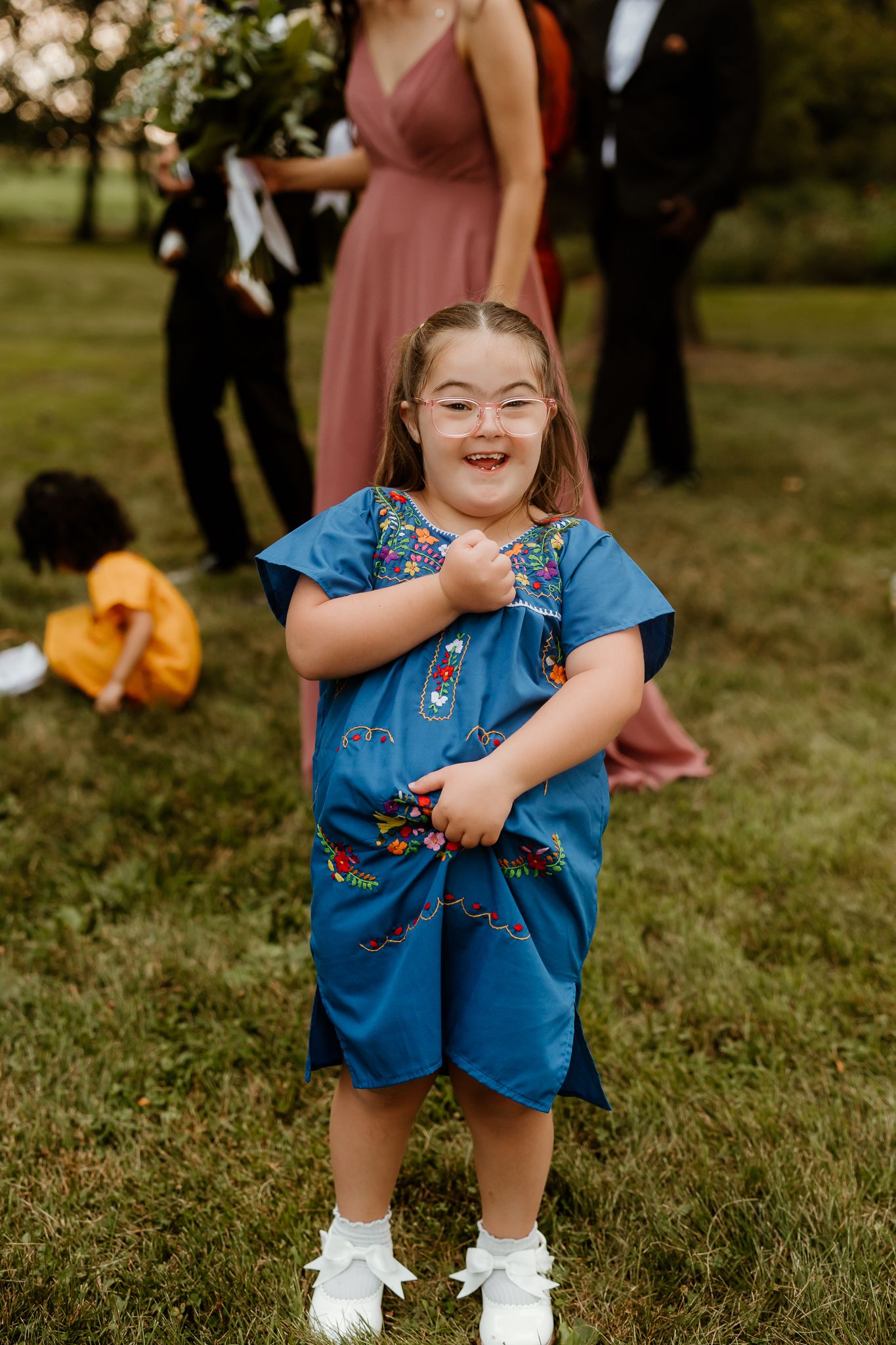 Young girl in blue embroidered dress smiling joyfully at outdoor wedding ceremony on green grass.