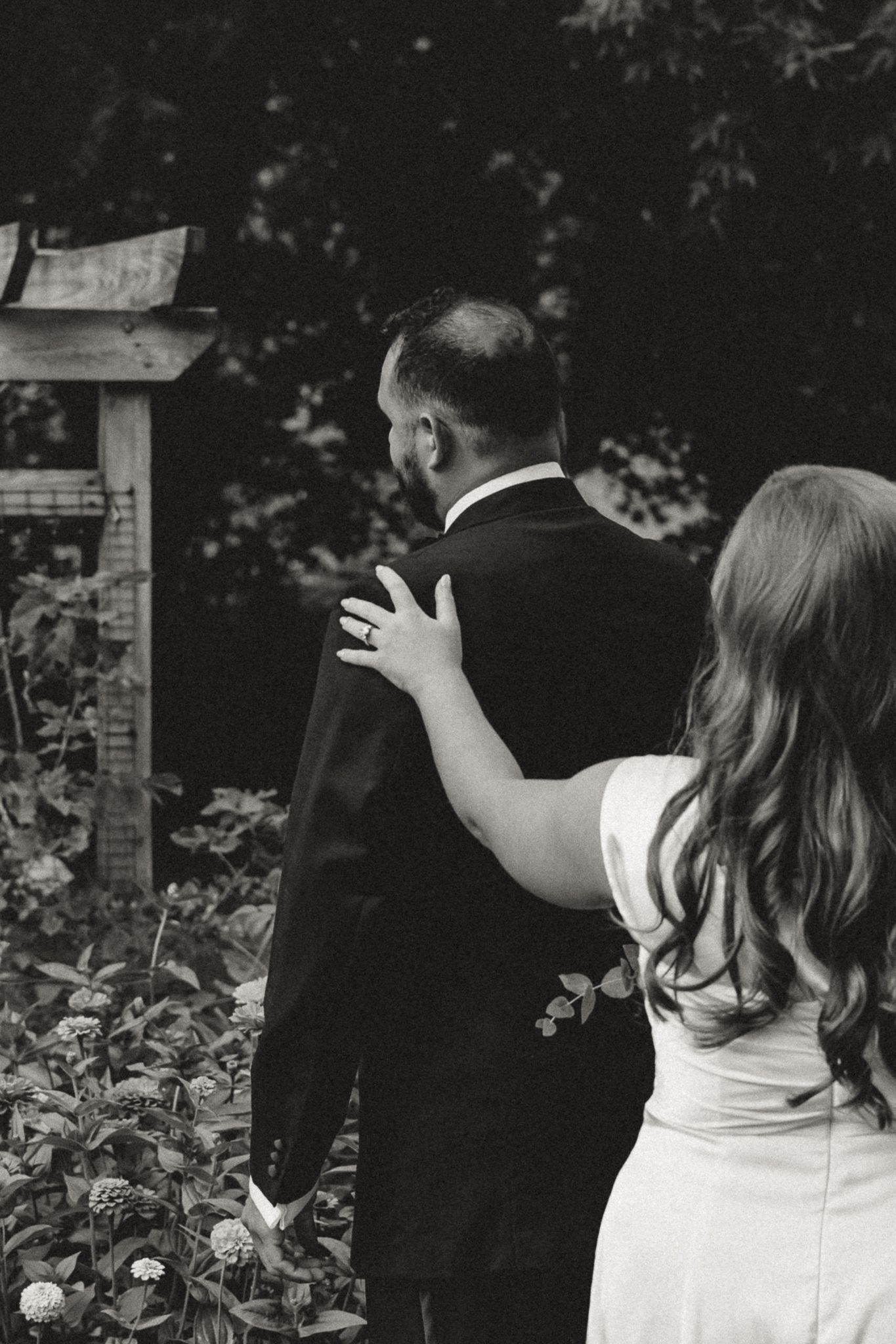 Black and white photo of a bride placing her hand on a groom's shoulder in a garden wedding setting.