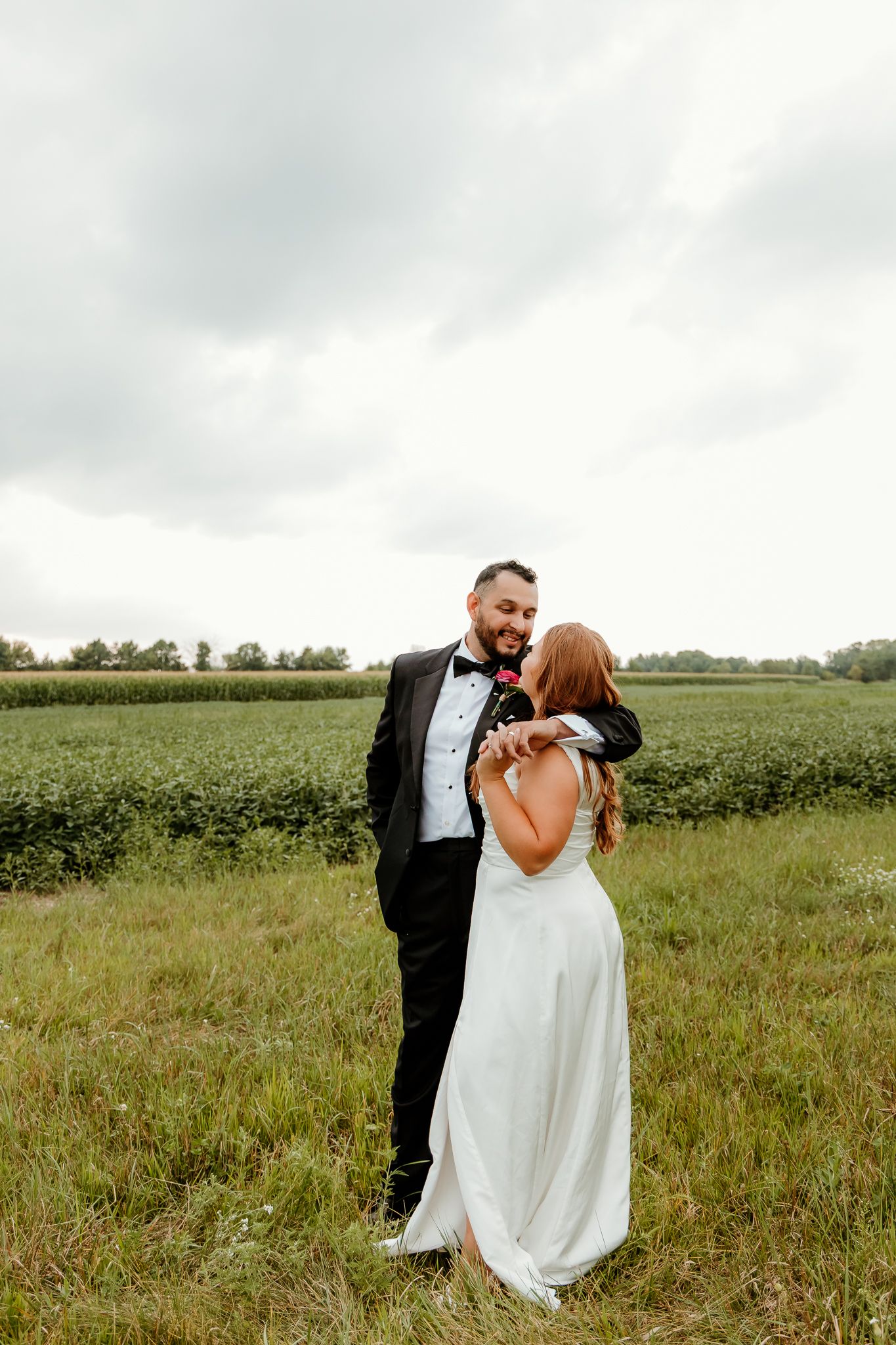 A bride and groom embrace in a lush green field, the groom in a black tuxedo and bride in a white gown, under a cloudy sky.