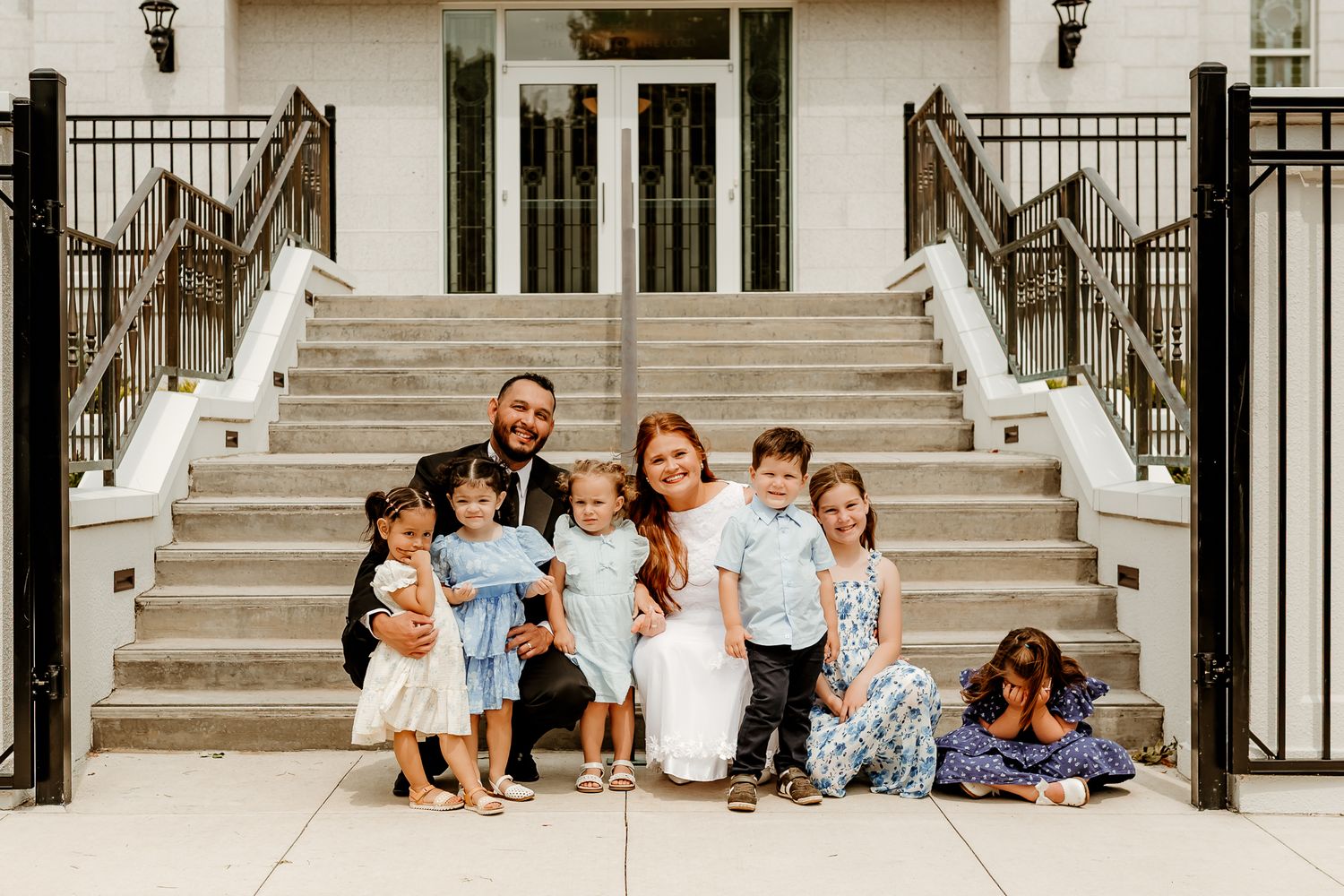 Family portrait with parents and five children posing on stone steps outside a white building.