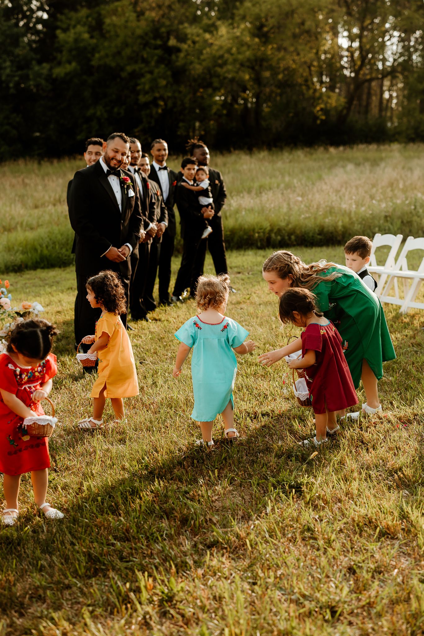 Children in colorful dresses walk down a grassy aisle as groomsmen in black tuxedos watch during an outdoor wedding ceremony.