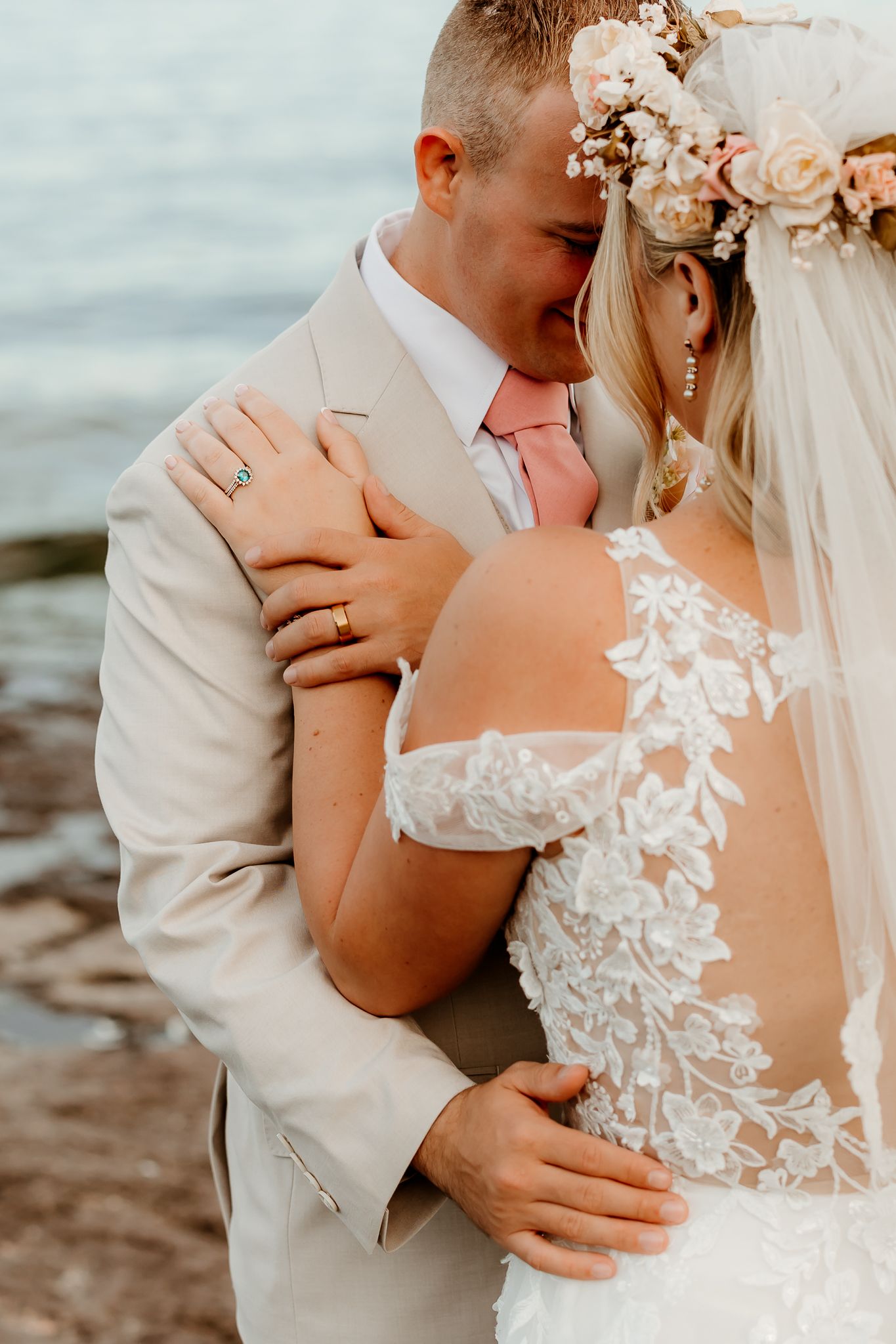 Bride and groom embrace near ocean, she wears lace off-shoulder gown and floral crown, he wears tan suit with pink tie.