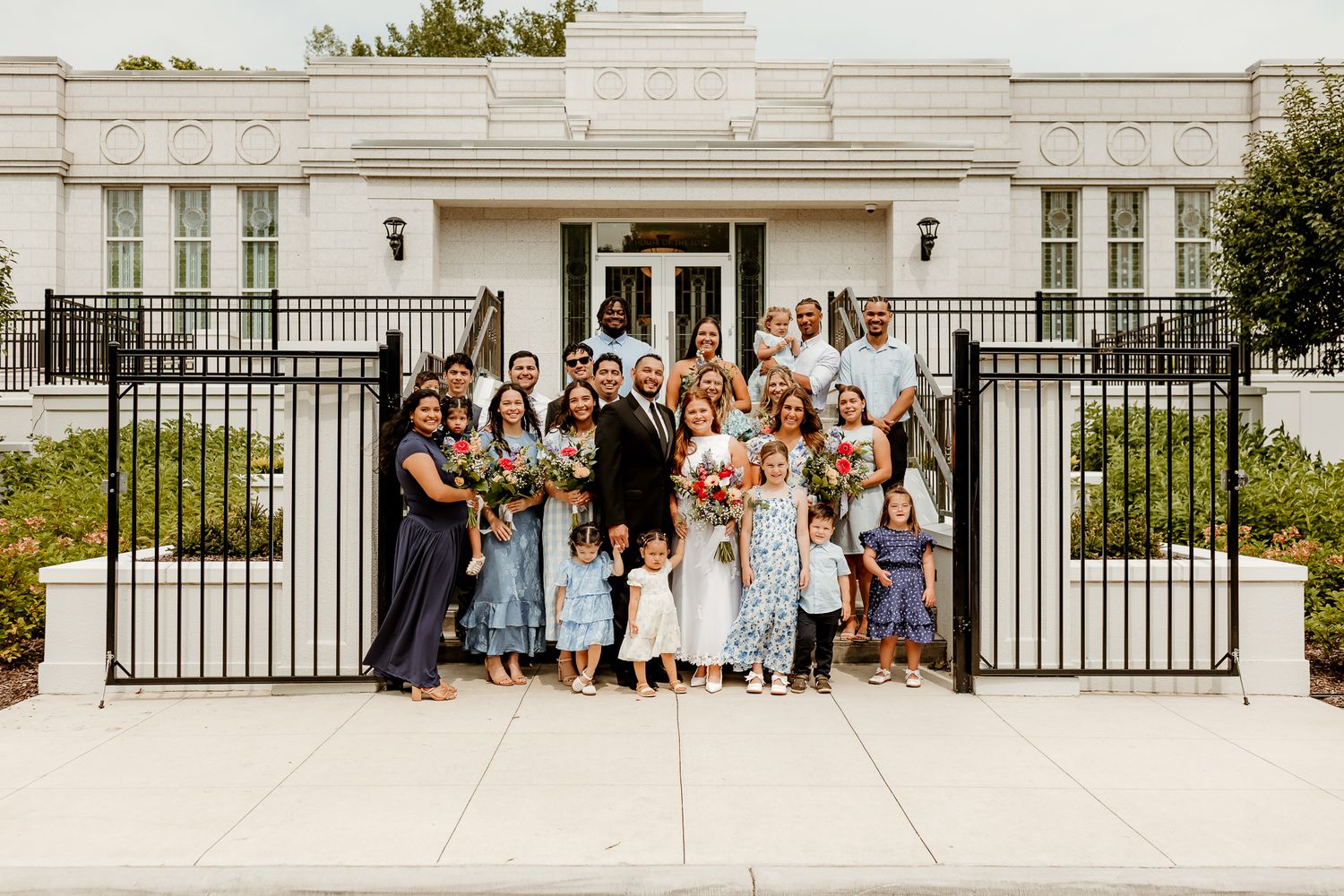 Large family group posing together outside a white building with iron gates, celebrating a wedding occasion.