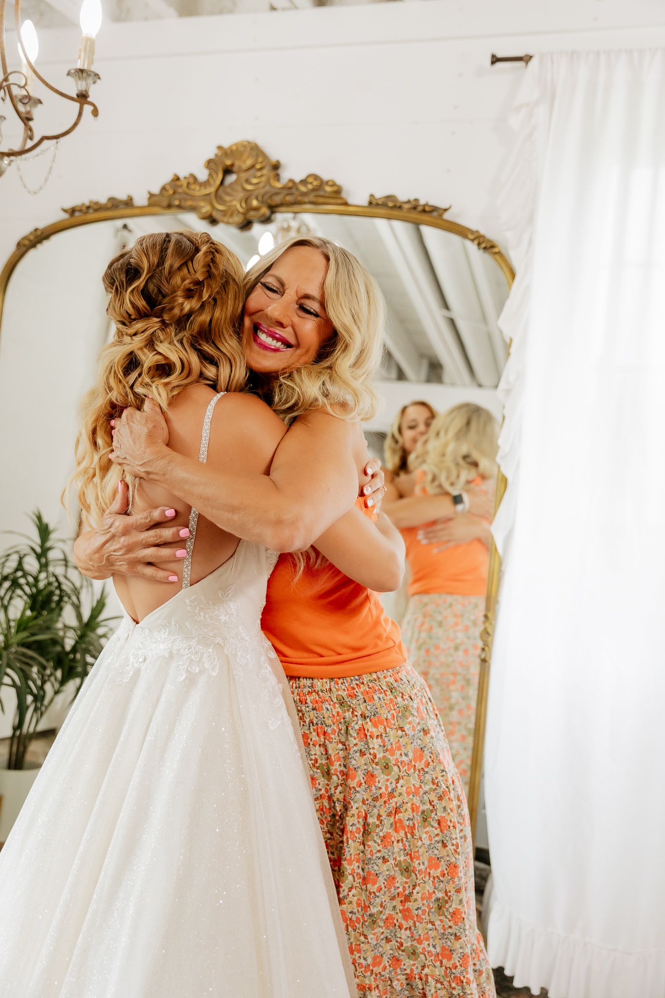 A bride in a white gown shares a joyful hug with a smiling woman in orange near a gold ornate mirror.