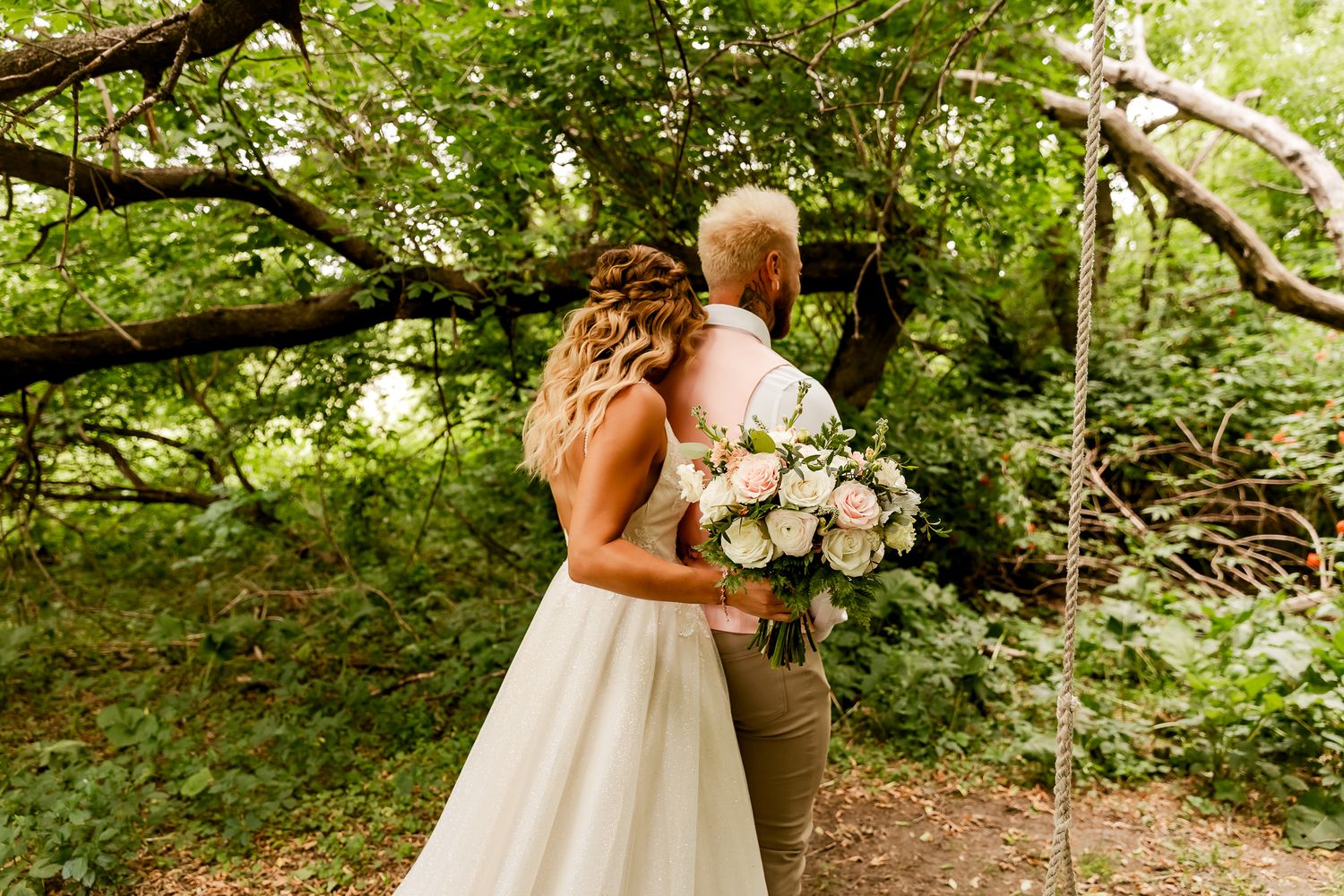Bride and groom stand back-to-back in lush green forest, bride holding elegant pink and white wedding bouquet.