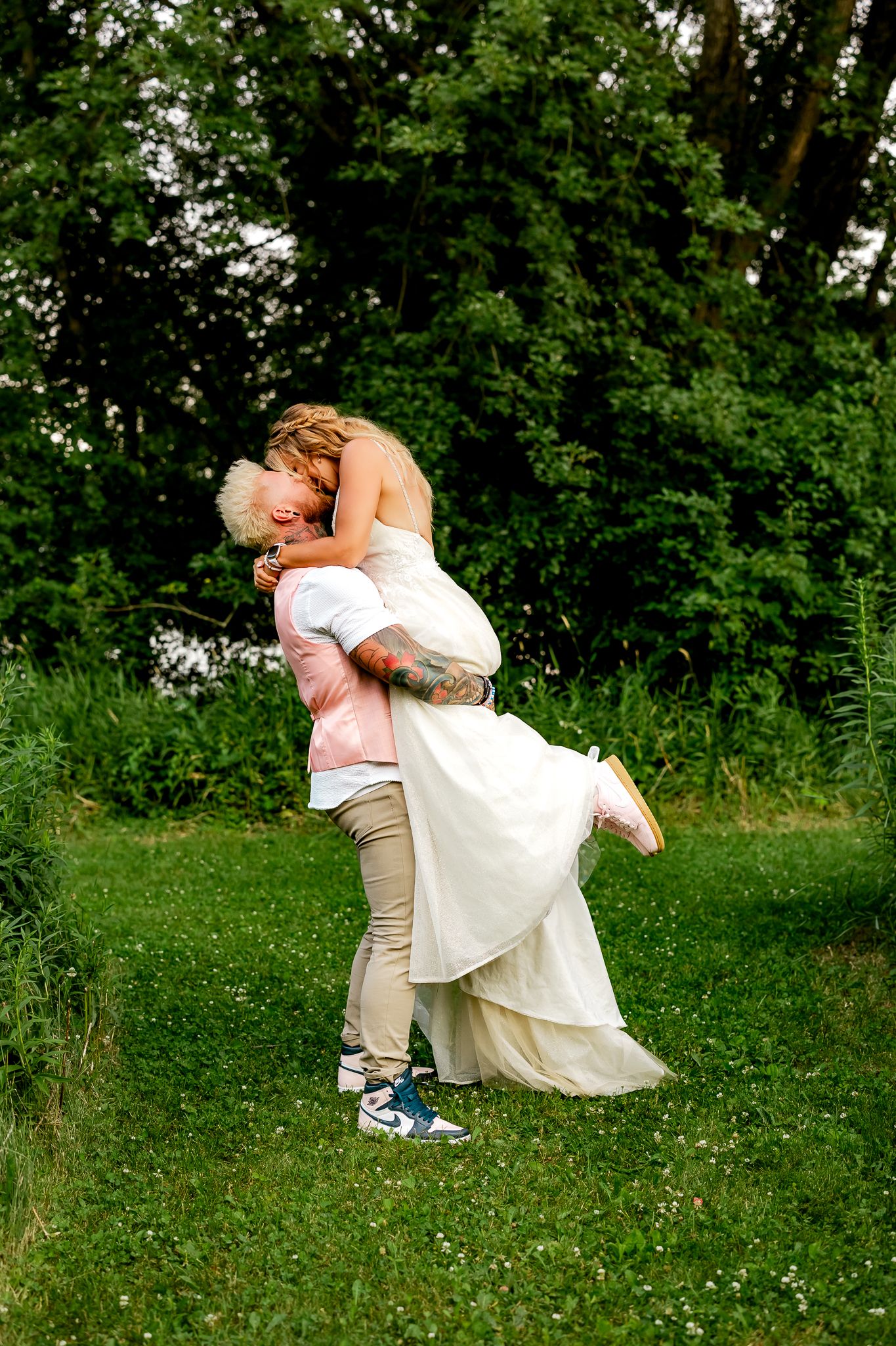 Groom lifts bride in white wedding dress outdoors on green grass, sharing a romantic kiss surrounded by lush trees.