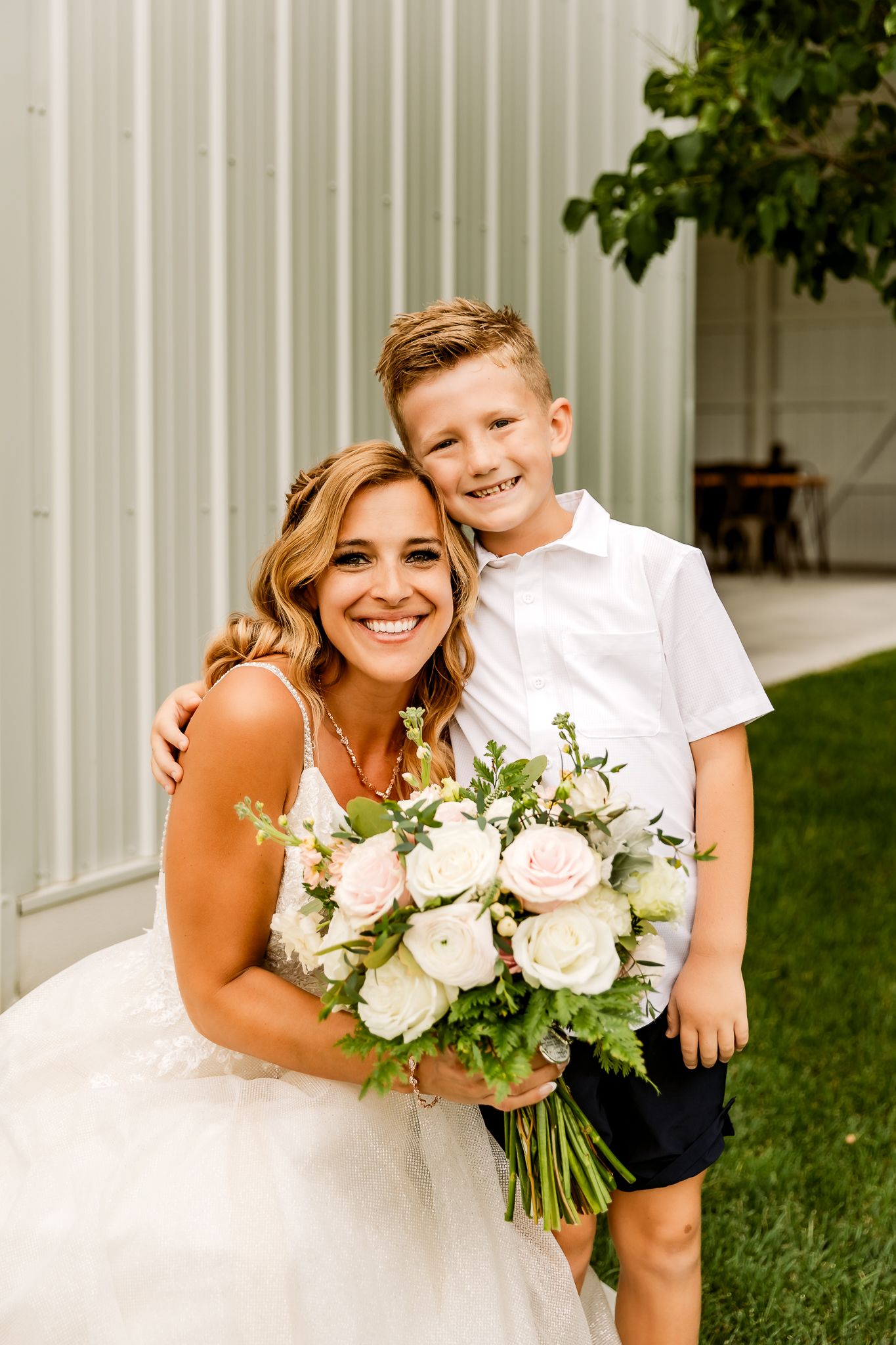 Smiling bride in white dress holding pink and white bouquet poses with young boy in white shirt outdoors.
