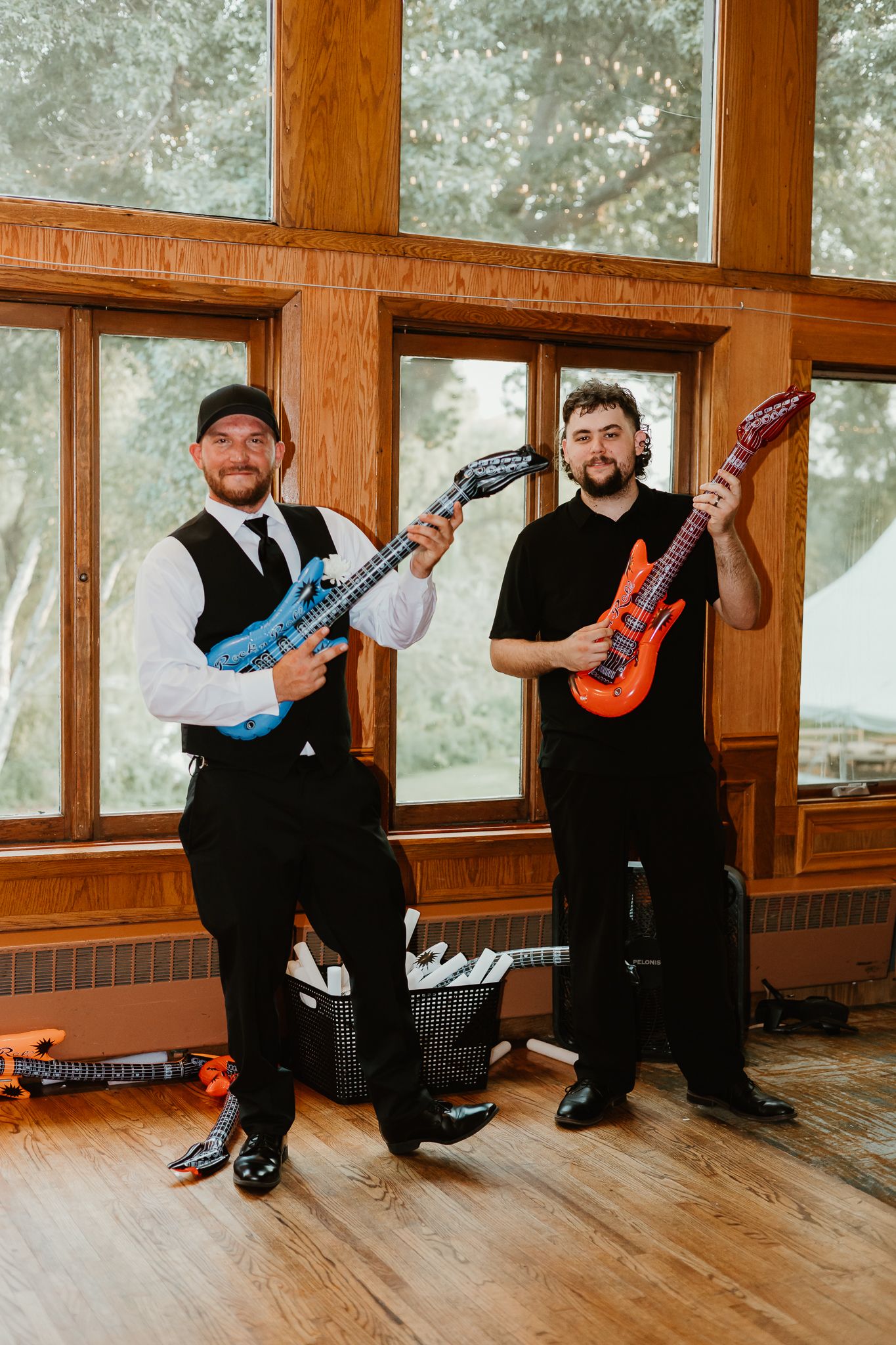 Two men in formal attire pose with colorful inflatable guitars in a wood-paneled room.