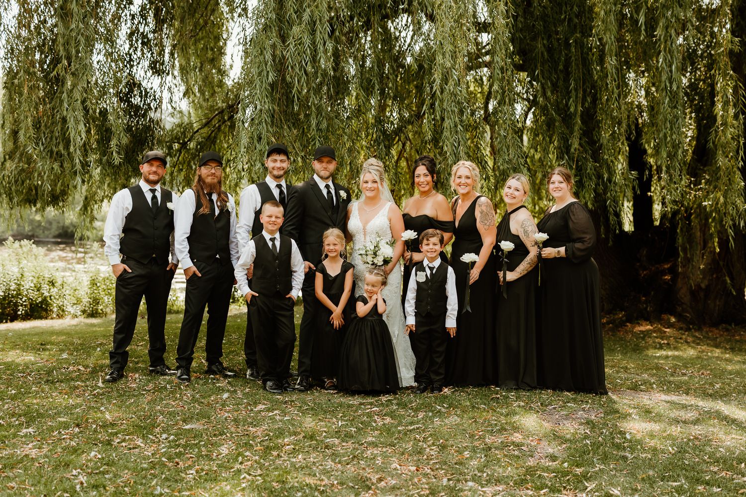 Wedding party in black attire poses under a weeping willow tree with bride and groom in white.