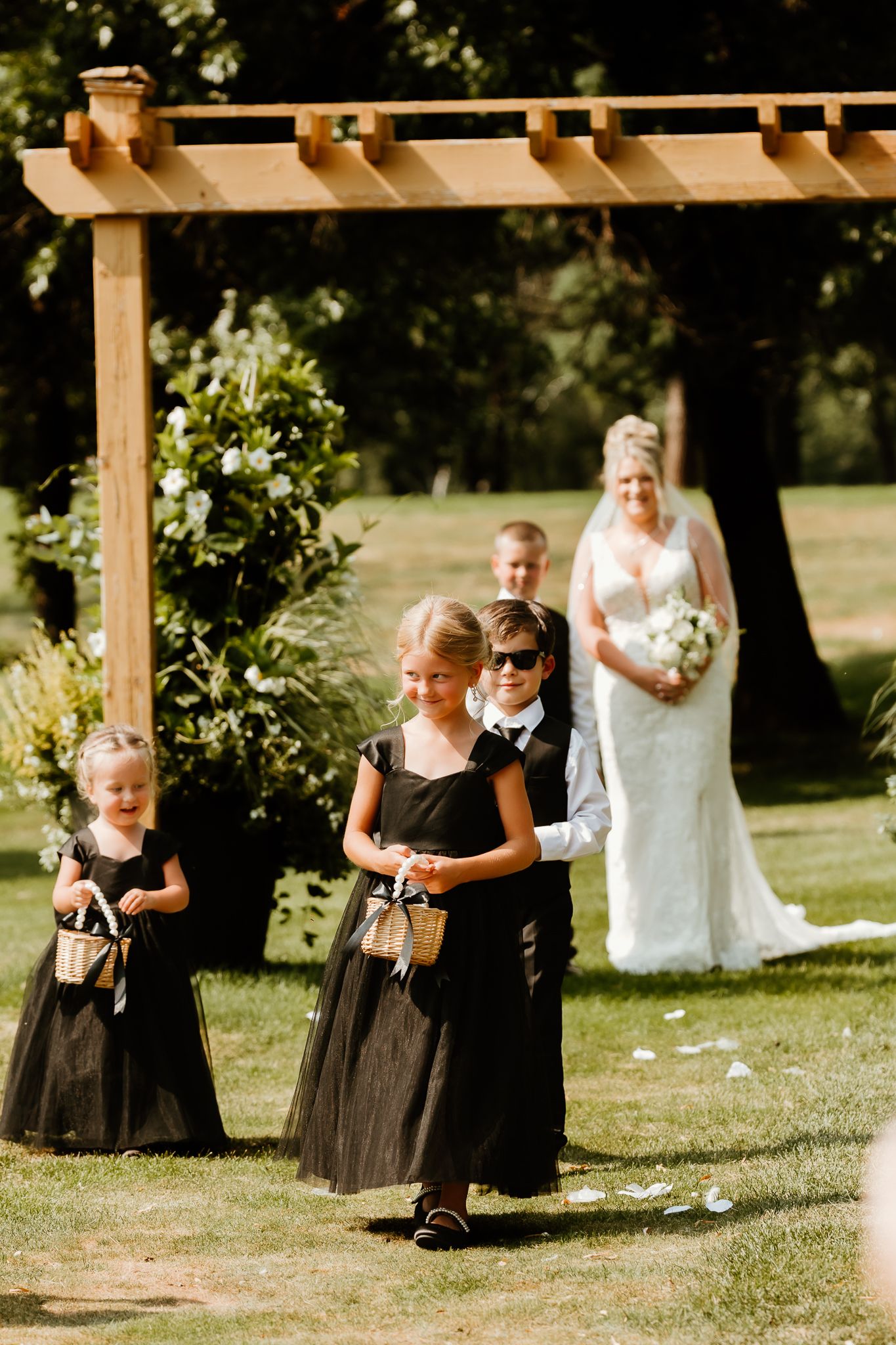 Flower girls in black dresses walk down the aisle as a bride in white holds bouquet at an outdoor wedding ceremony.