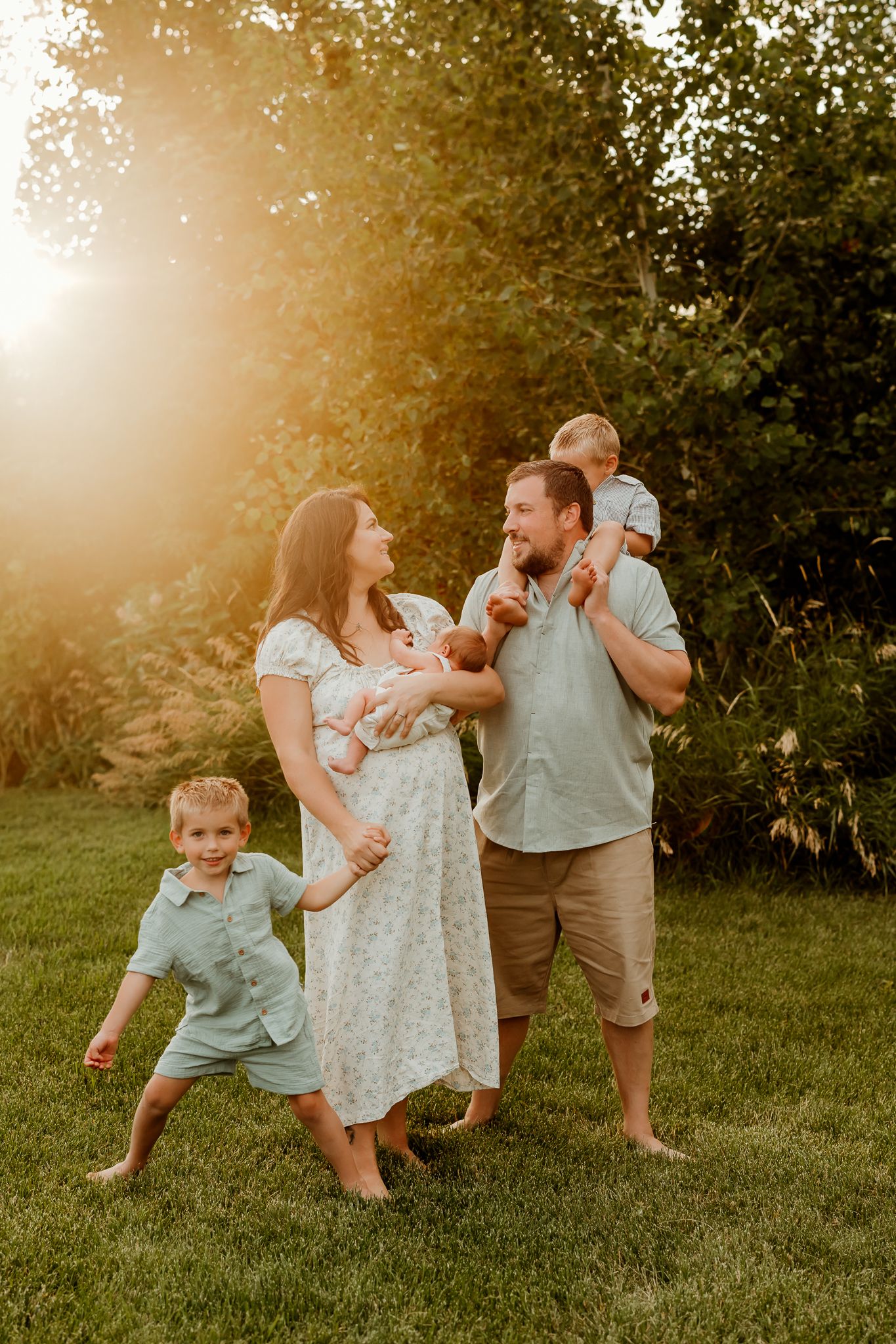 Family of five poses outdoors at golden hour, parents holding children on lush green grass with warm sunlight.