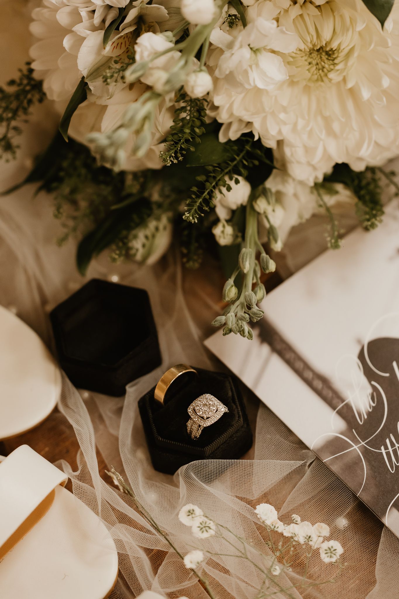 A diamond engagement ring in a black velvet box surrounded by white floral bouquet, sheer veil, and elegant stationery.