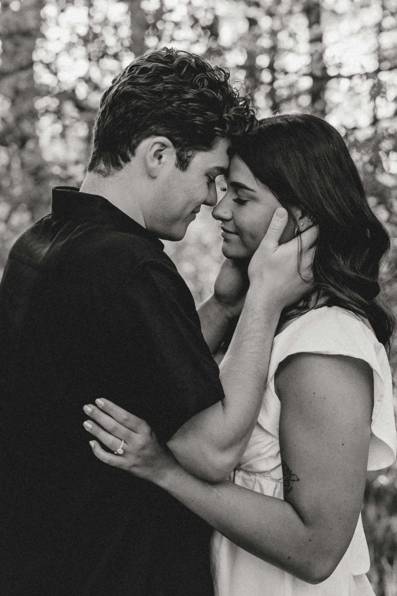 Black and white romantic portrait of couple touching foreheads tenderly in forest setting.
