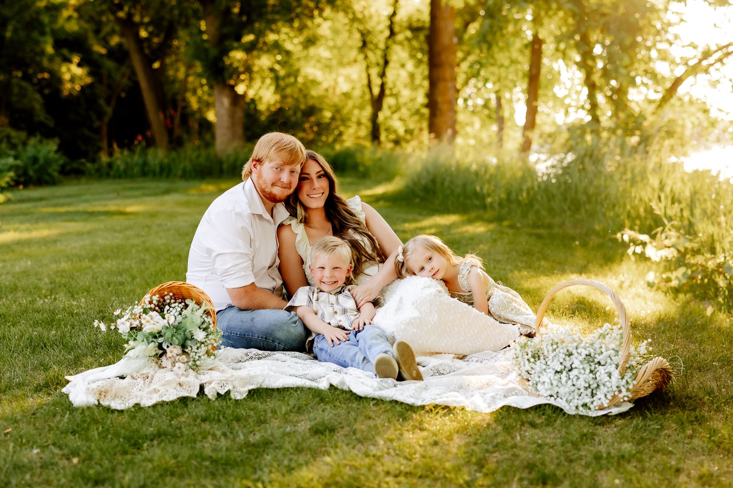 Happy family of four with two toddlers sitting on a blanket in a sunny green park with flowers.