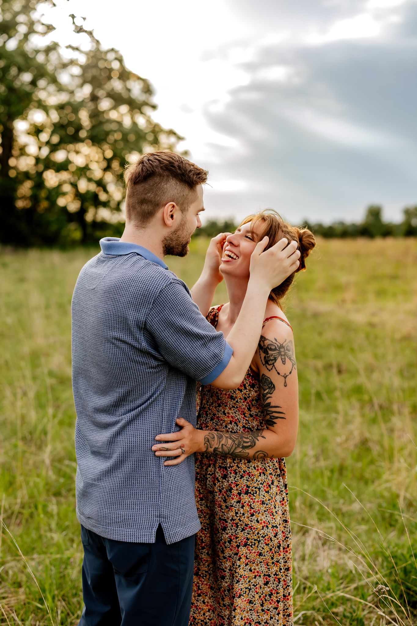 A loving couple embraces in a sunlit meadow, the woman in a floral dress with tattoos smiling warmly.