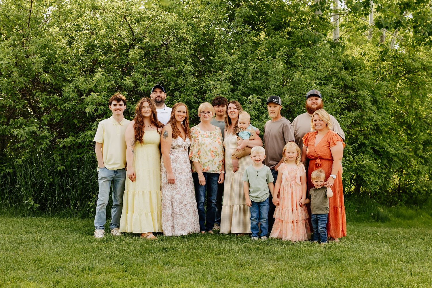 Extended family posing together on green lawn with dense foliage background during outdoor photo session.