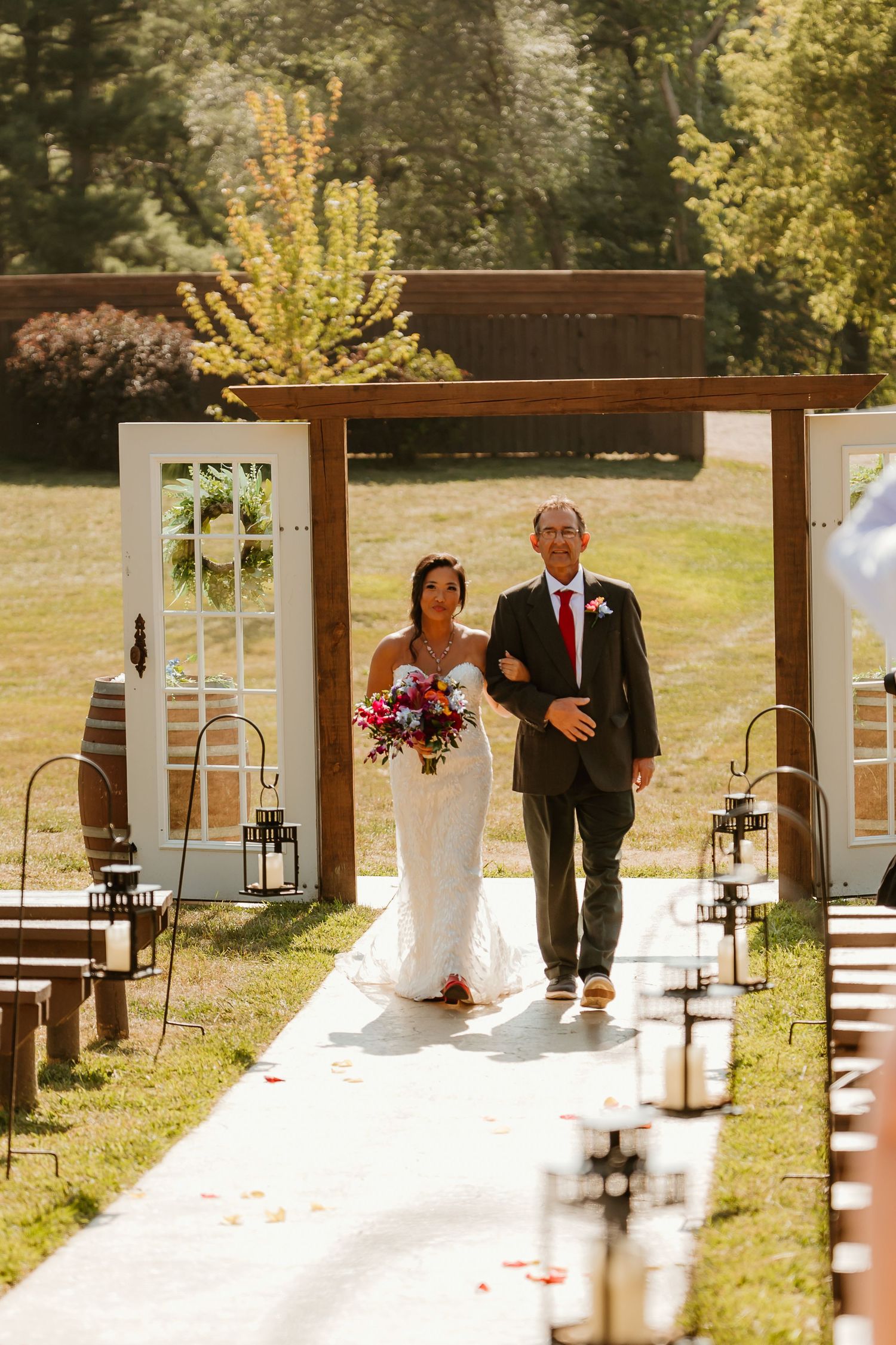 Bride walks down outdoor aisle with escort past white doors and lanterns at a rustic garden wedding ceremony.