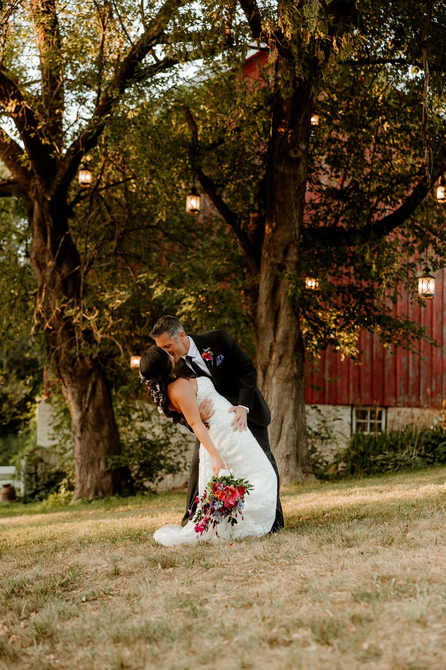 Bride and groom share a romantic dip kiss outdoors near a red barn with string lights hanging from trees at golden hour.