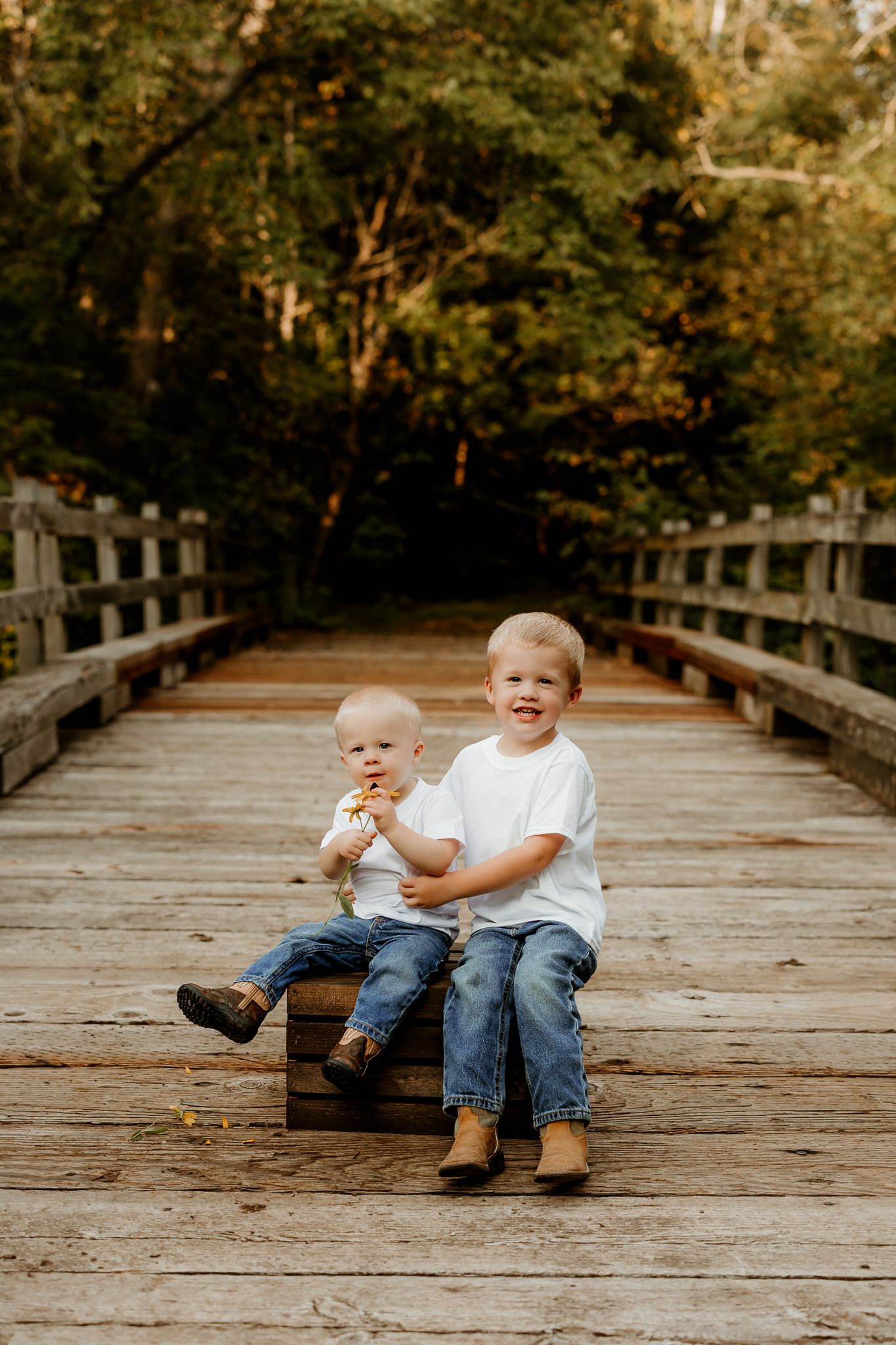 Two young boys in white shirts and jeans sit smiling on a wooden bridge surrounded by lush green trees.