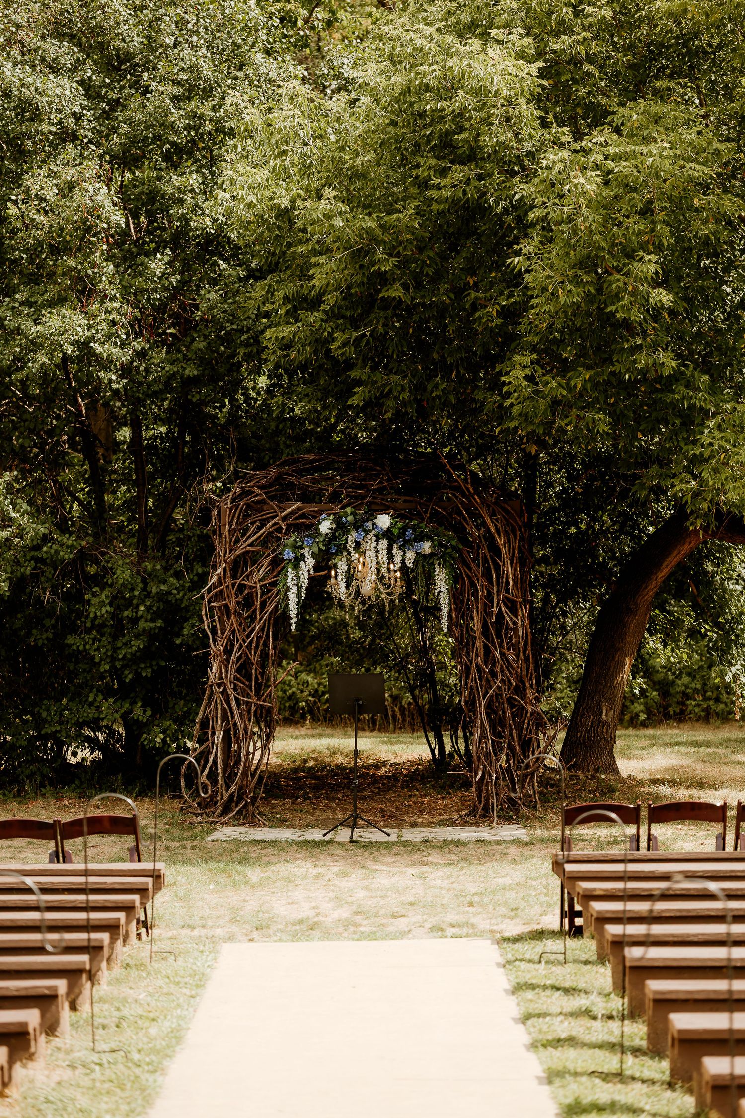 Rustic twig arch wedding ceremony setup with wooden chairs lining white aisle path surrounded by lush green trees.