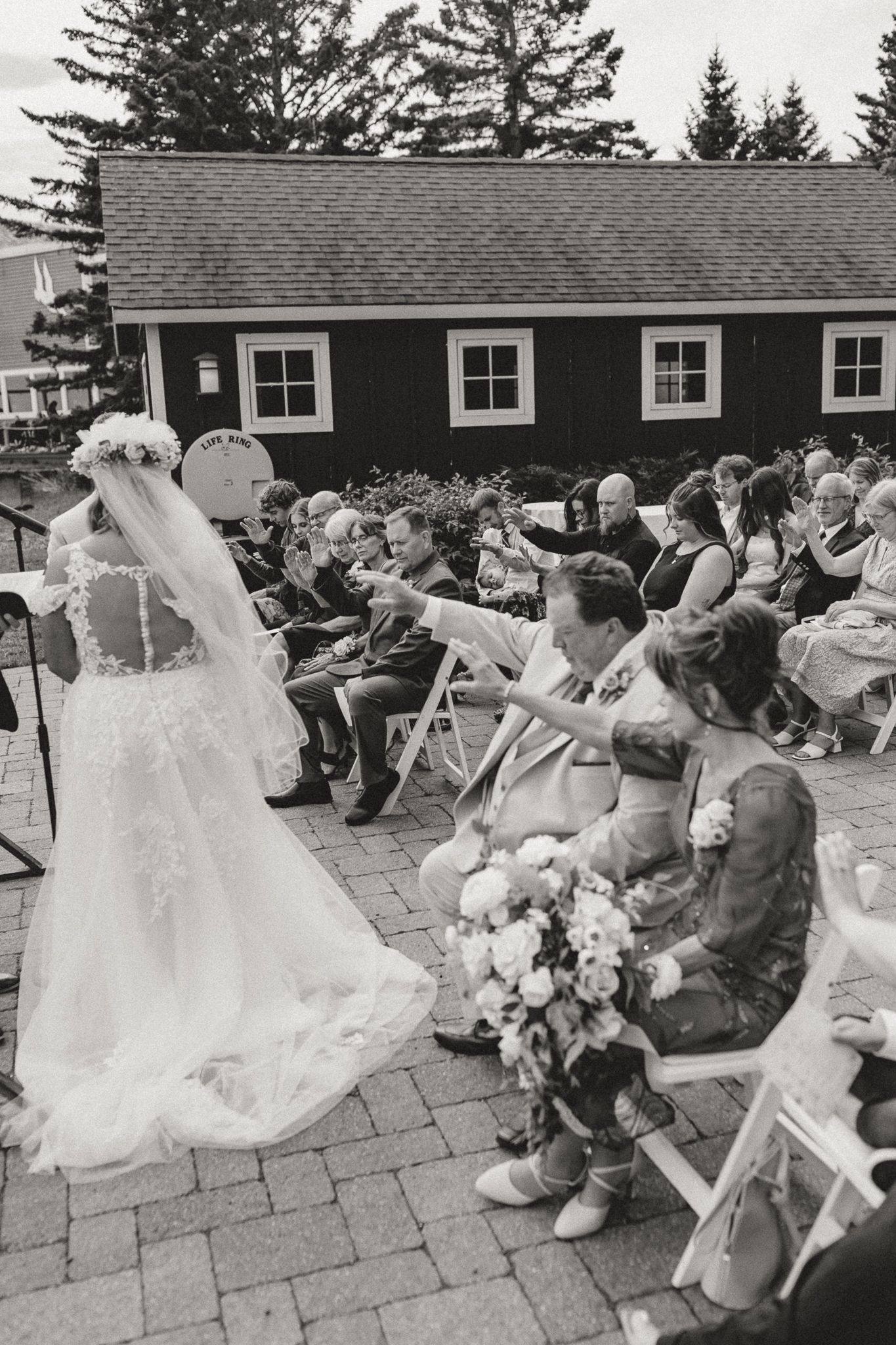 Black and white outdoor wedding ceremony on brick patio with bride in lace gown and veil, guests seated in white chairs.