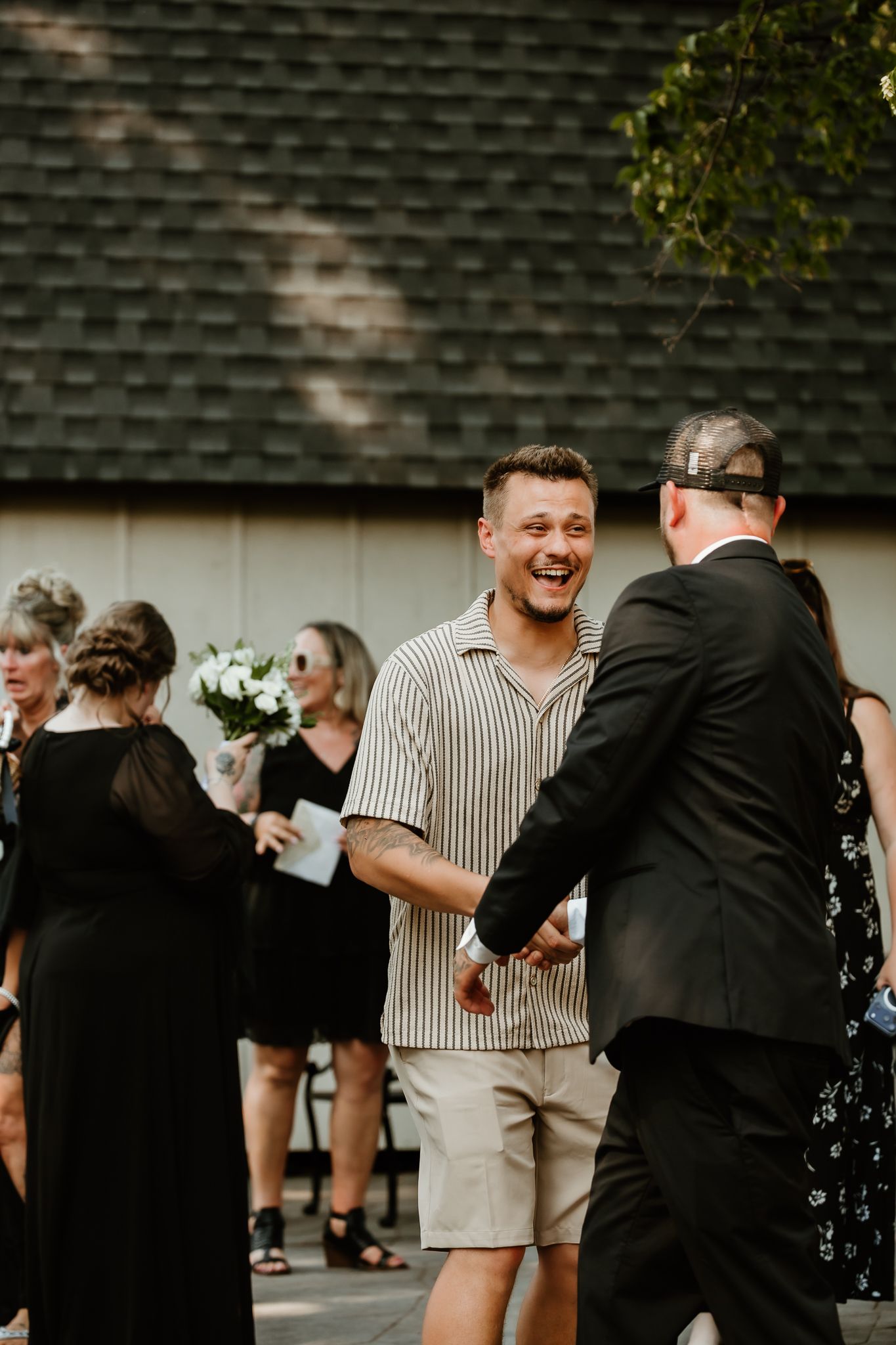 Man in striped shirt laughing and shaking hands at an outdoor wedding celebration with guests in black attire.