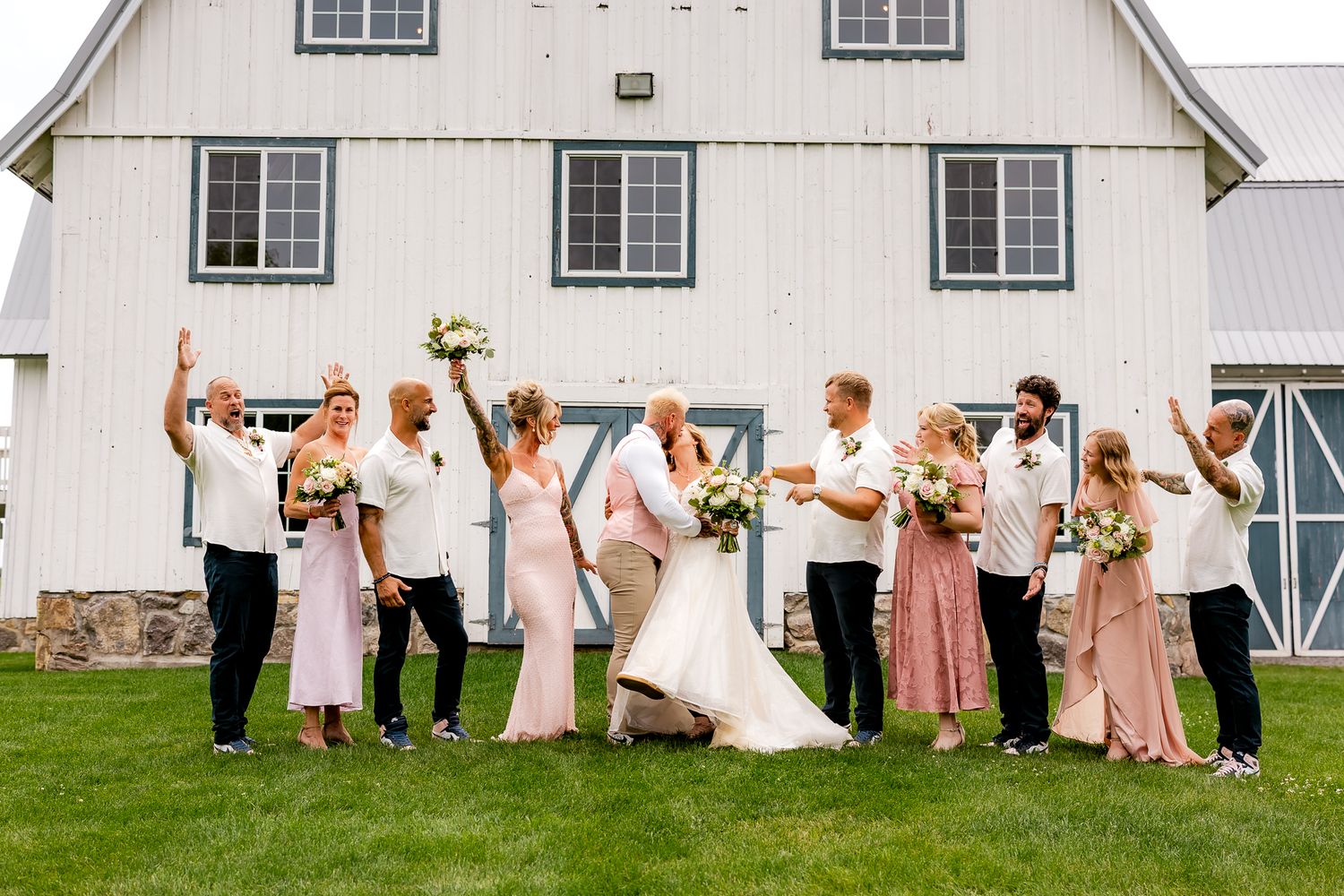 Wedding party celebrating in front of a white barn, bride and groom surrounded by bridesmaids and groomsmen cheering.